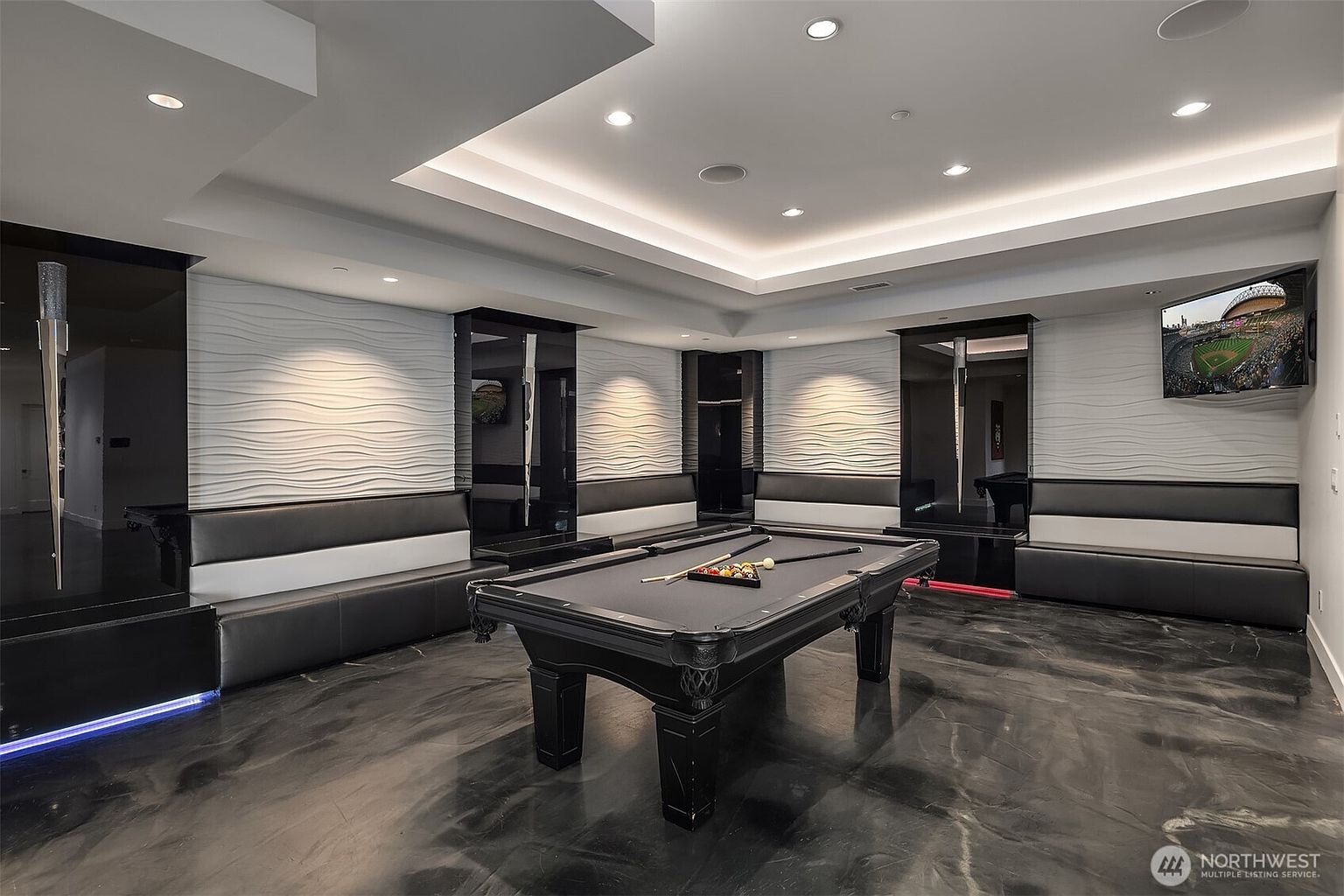 This modern, high-end game room features a central pool table set against a backdrop of textured, wave-patterned wall panels and sleek black built-in bench seating. The room is illuminated by recessed lighting within a tray ceiling, while a wall-mounted television displays a sports broadcast, enhancing the entertainment-focused atmosphere. The polished, dark reflective flooring adds a sophisticated, contemporary aesthetic to this luxurious recreational space.