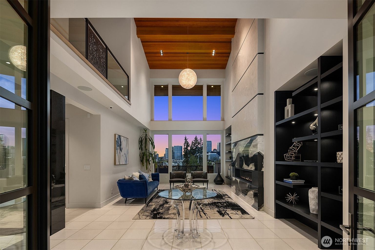 This expansive, double-height living room features a dramatic wood-paneled ceiling and a striking, full-height stone fireplace surround with a dark, artistic mantle. A glass-topped circular table sits in the foreground, while a plush blue sofa and two leather armchairs provide comfortable seating near large windows that frame a city skyline view. The space is defined by its open, airy atmosphere, polished light-colored tile flooring, and a sleek, dark built-in shelving unit that adds a sophisticated contrast to the bright, modern interior.