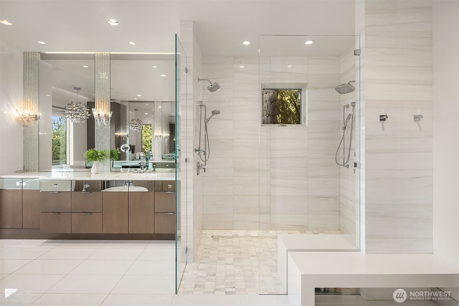 This luxurious primary bathroom features a spacious walk-in shower enclosed by glass panels, showcasing light-toned wall tiles and a mosaic-tiled floor. Adjacent to the shower is a sleek vanity with dark cabinetry, a large mirror, and modern lighting fixtures. The room is bright and contemporary, with a clean aesthetic that emphasizes high-end finishes and an open, airy layout.