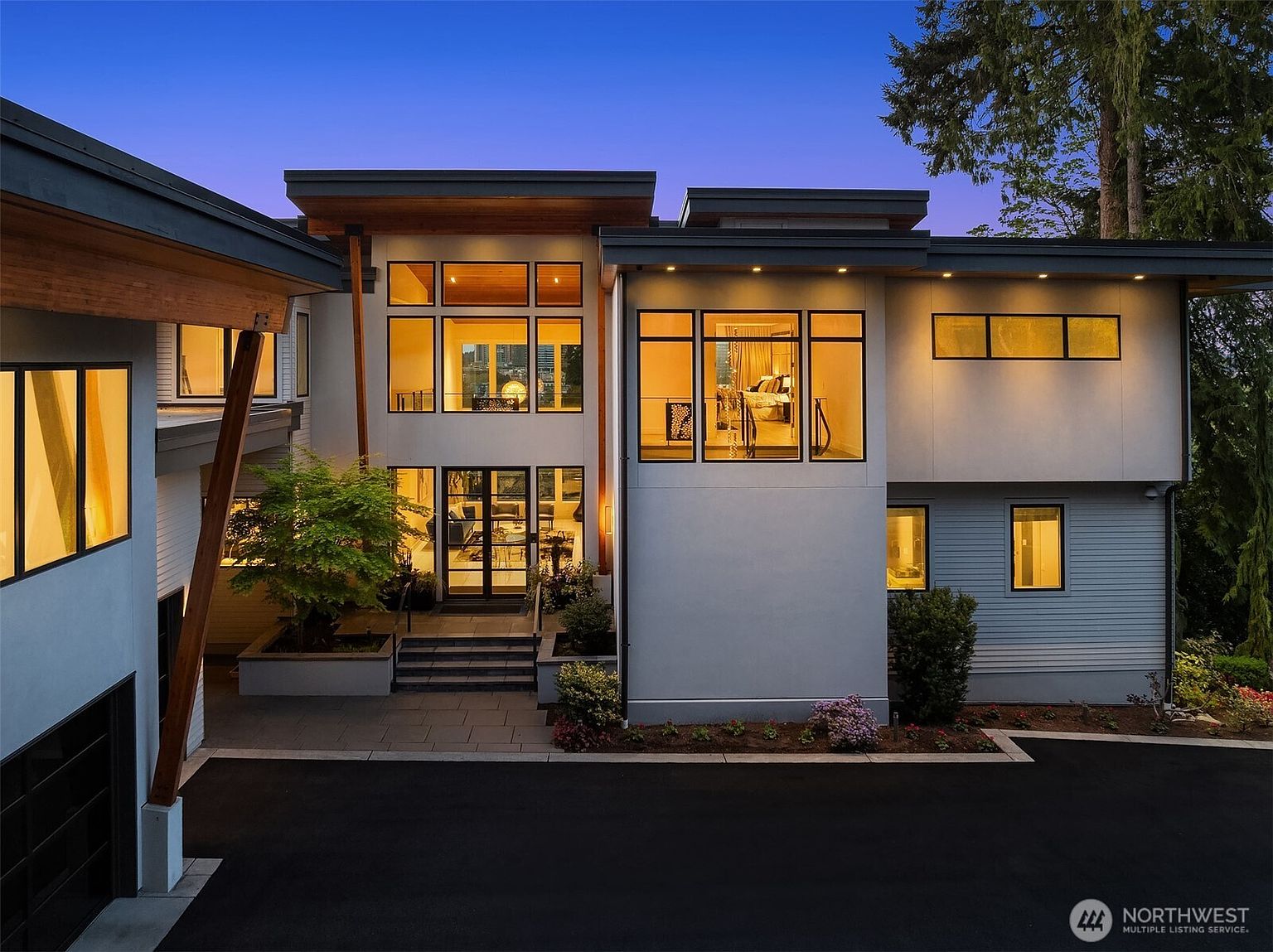 This modern architectural residence features a striking multi-level facade with a mix of smooth light-toned surfaces and horizontal siding. The home is defined by its expansive glass windows, including a prominent central entryway with a tall glass door system and a second-story bedroom visible through large windows. A paved driveway leads to the front entrance, which is accented by a mature tree and clean, minimalist landscaping.