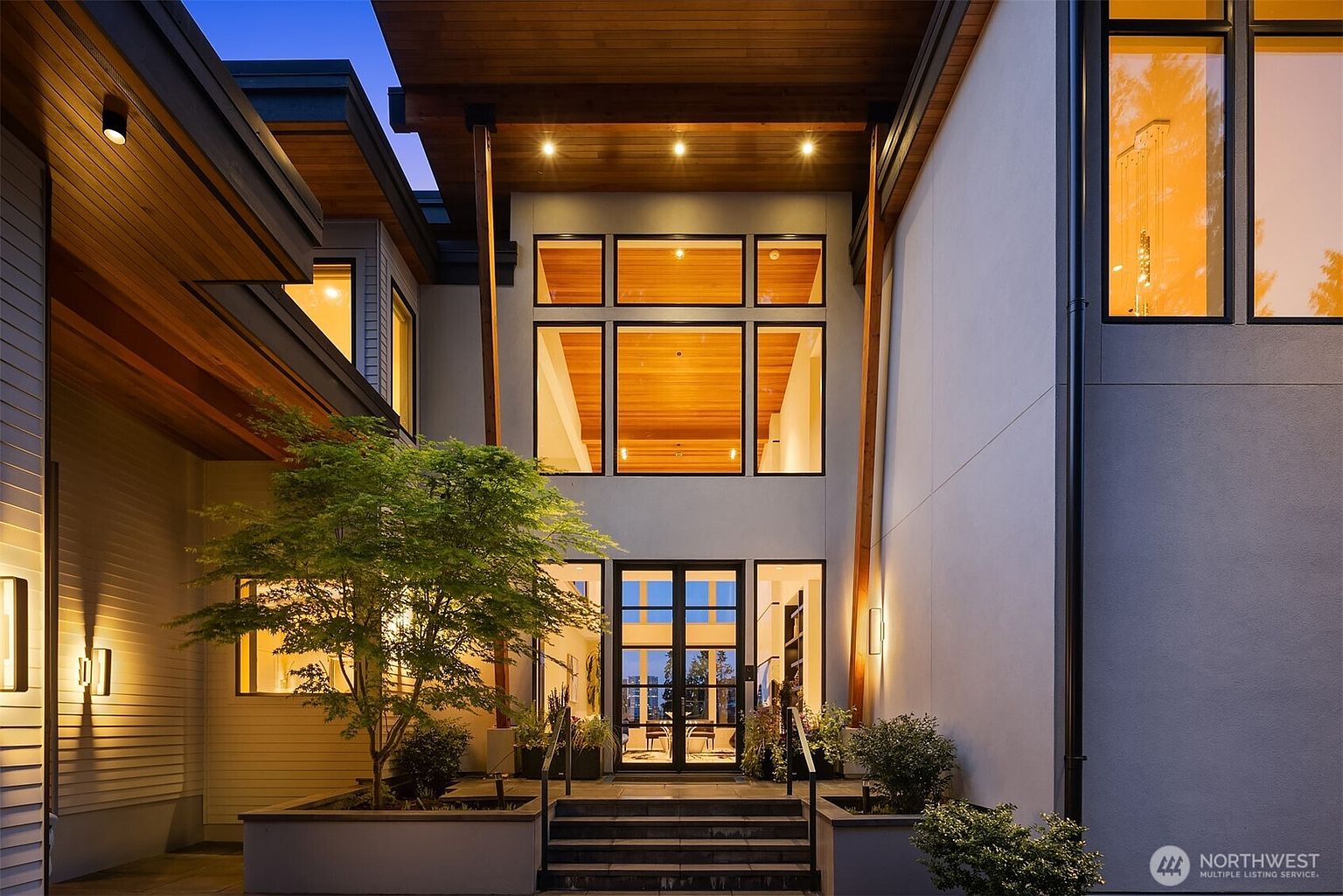 This striking modern entryway features a grand, double-height glass facade framed by warm wood accents and clean, light-colored walls. A set of stone steps leads up to a central glass-paned door, flanked by symmetrical planters and a manicured tree, all illuminated by soft, warm exterior lighting. The architectural design emphasizes clean lines and a welcoming, sophisticated atmosphere at dusk.