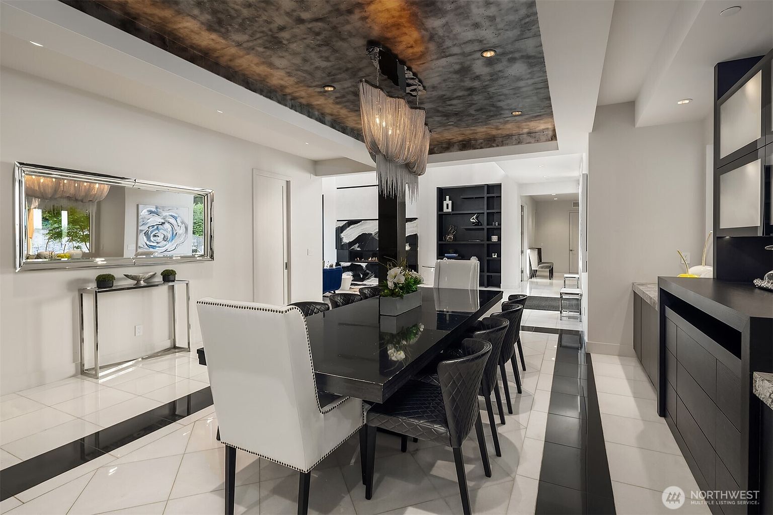 This sophisticated dining room features a high-gloss black table surrounded by white upholstered chairs, set against a backdrop of white walls and a striking metallic-finished ceiling. A large, modern chandelier with cascading elements serves as a focal point above the table, while a wide mirror reflects the adjacent living area. The space is defined by a clean, high-contrast aesthetic with polished white flooring accented by dark border inlays.