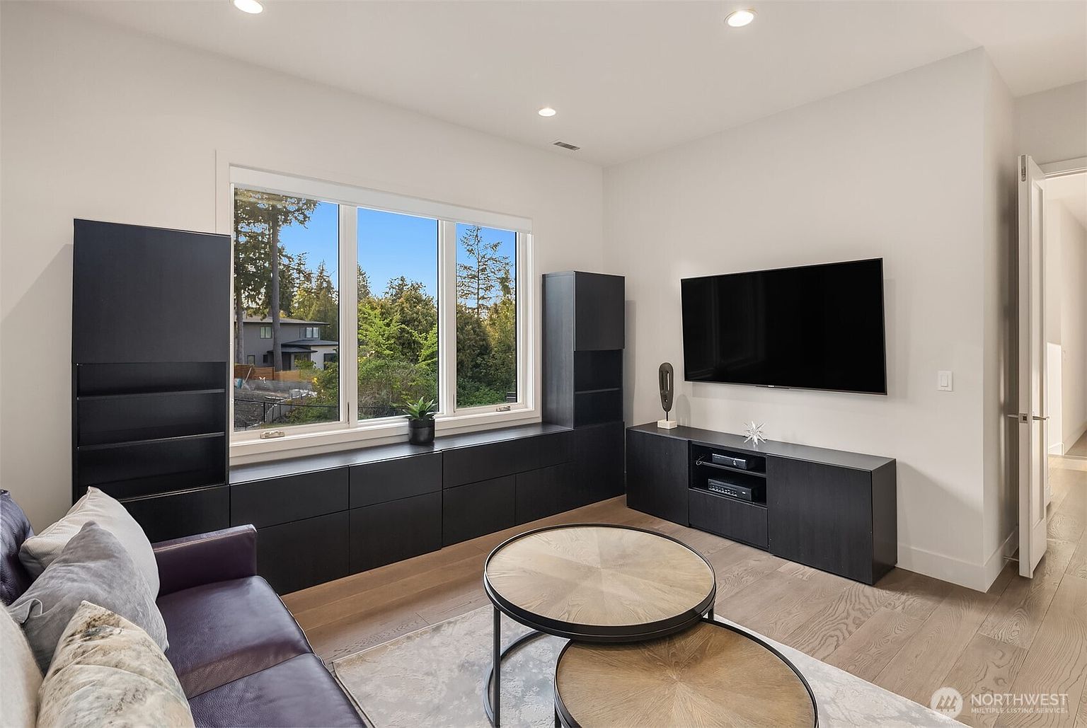 This contemporary living room features a sleek, dark-toned built-in cabinetry unit that spans the wall beneath a large window, providing ample storage and display space. A wall-mounted television sits above a matching media console, creating a clean and minimalist entertainment area. The room is furnished with a dark leather sofa and a pair of round, light wood coffee tables, all set upon a neutral area rug over light-toned wood flooring.