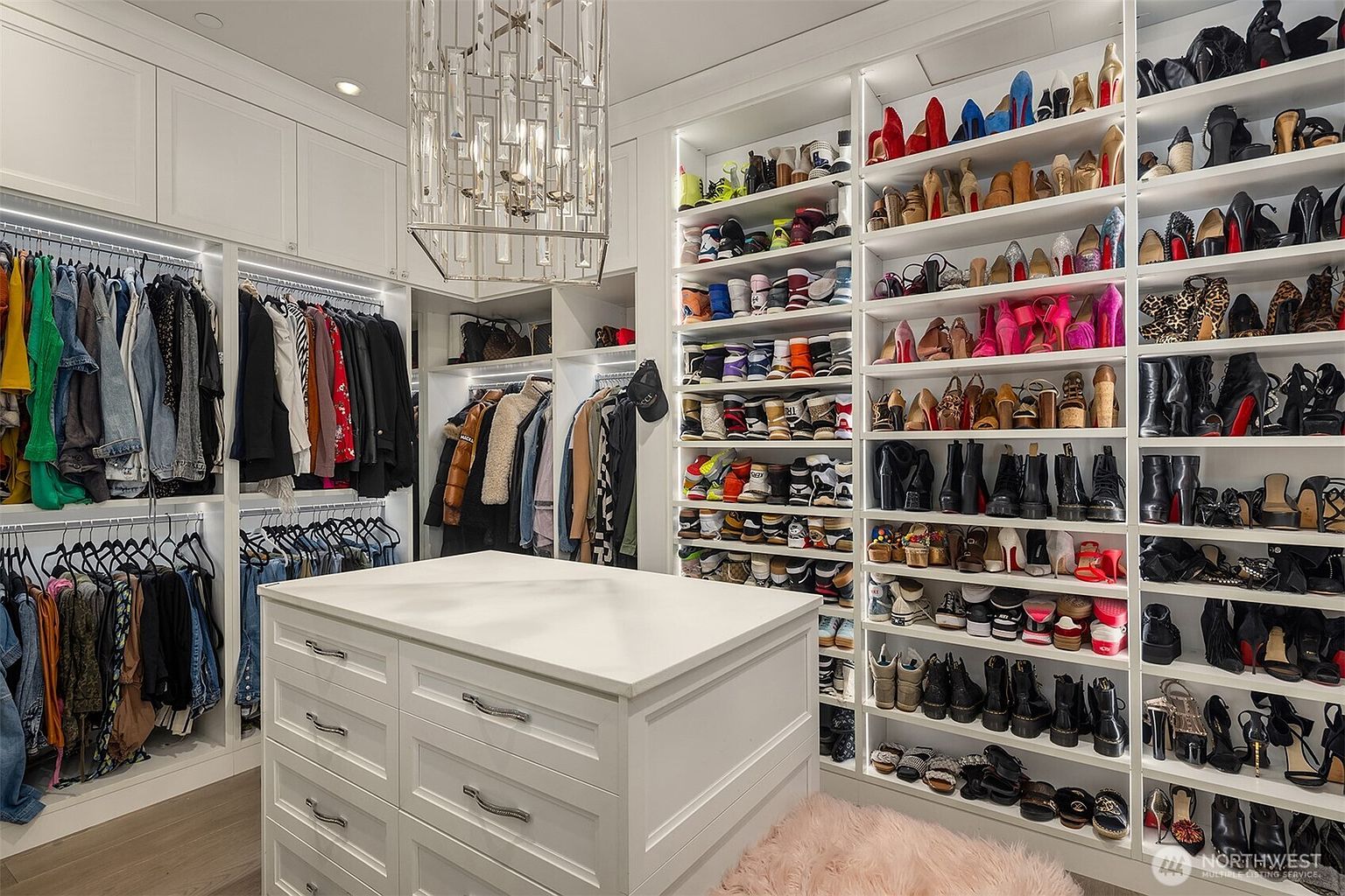 This luxurious walk-in closet features extensive custom white cabinetry with integrated LED lighting, providing ample storage for clothing and an impressive collection of footwear. A central island with drawers offers additional organization, while a sophisticated crystal chandelier serves as a focal point above. The space is finished with light-toned flooring and a plush pink accent stool, creating an elegant and highly organized dressing area.