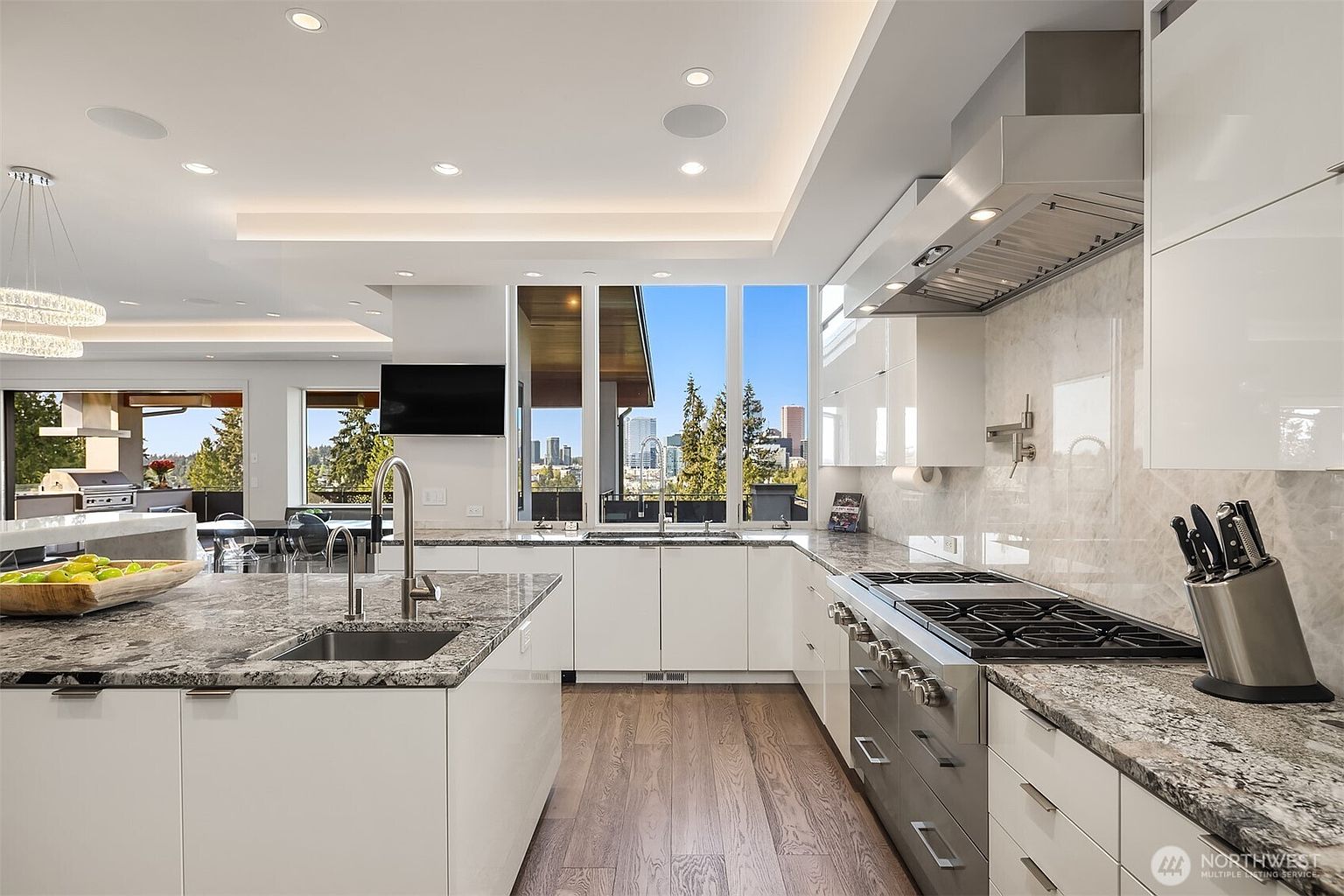This modern, open-concept kitchen features sleek white cabinetry, a large central island with a stone countertop, and high-end stainless steel appliances. Large windows provide a clear view of the city skyline, while recessed lighting and a contemporary chandelier create a bright, airy atmosphere. The space is finished with light wood flooring, offering a sophisticated and clean aesthetic suitable for high-end living.