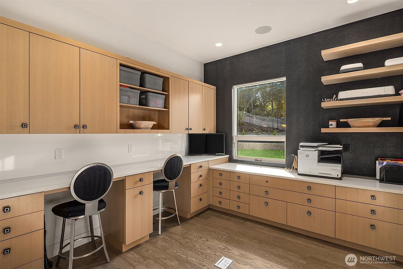 This functional home office features extensive light wood cabinetry and a long, continuous desk surface that provides ample workspace. The room is accented by a dark, textured feature wall and open shelving, creating a modern and organized aesthetic. A window above the desk allows natural light to brighten the space, which is furnished with two sleek, circular-backed stools.