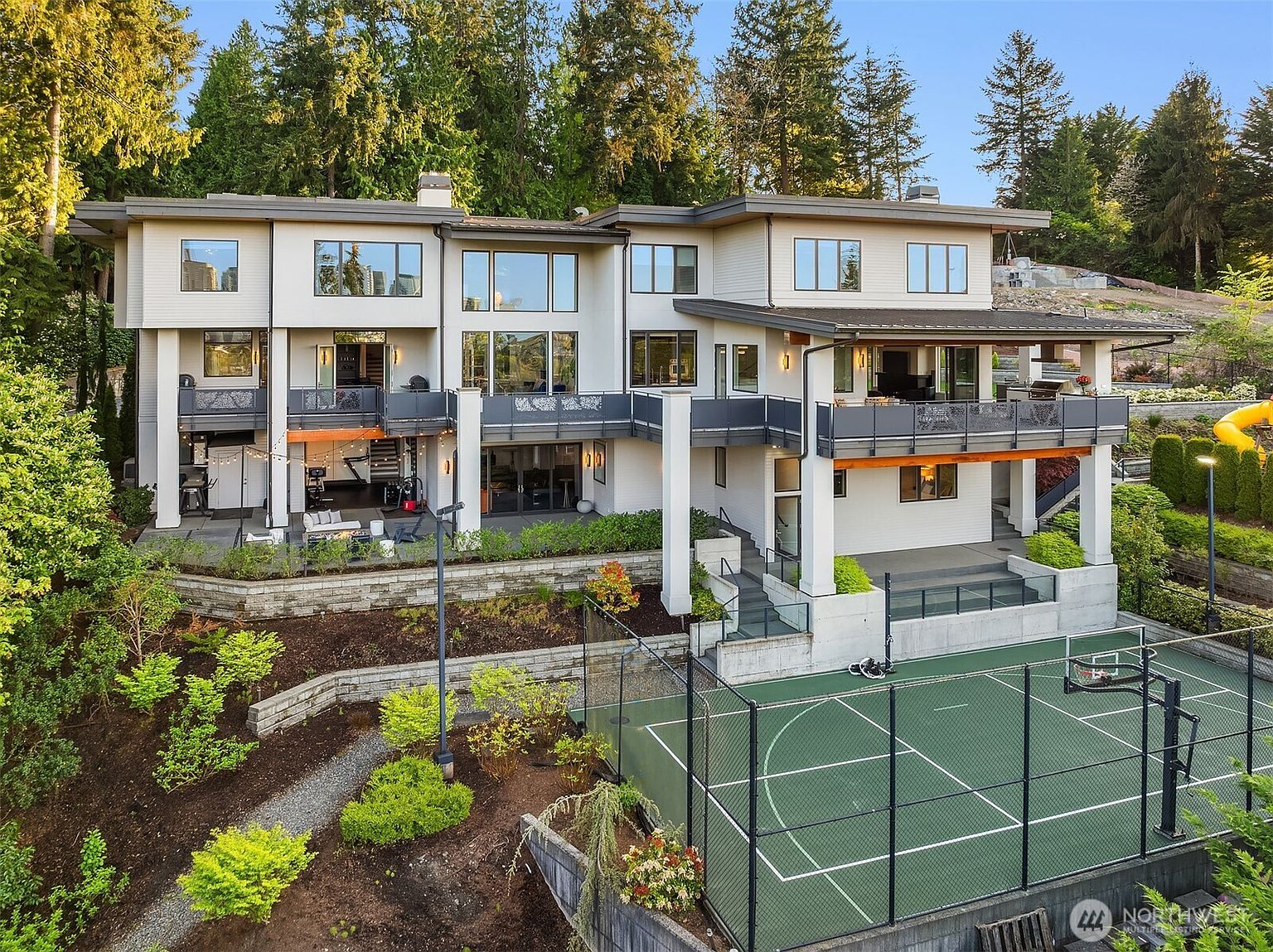 This expansive multi-level contemporary residence features a light-colored exterior with clean lines and large windows that offer a seamless connection to the outdoors. The rear of the home showcases multiple levels of balconies and a covered patio area, overlooking a private sports court and tiered landscaping. The elevated perspective captures the home's integration into the sloped, wooded lot, emphasizing its luxury design and recreational amenities.