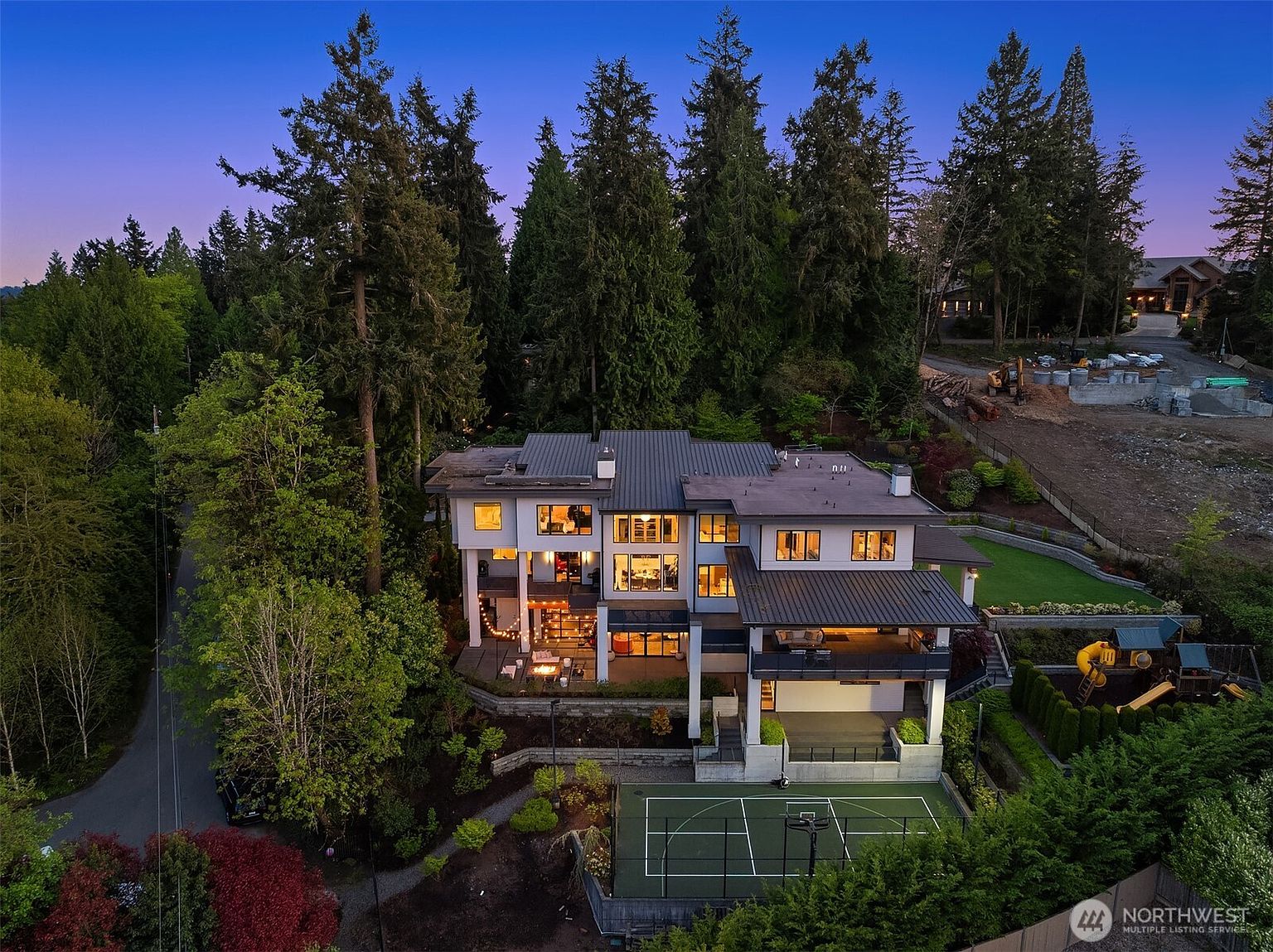 This high-angle aerial view captures a large, contemporary multi-story residence nestled against a dense forest backdrop during the twilight hour. The home features a dark metal roof, expansive windows, and a multi-level rear deck with integrated lighting, overlooking a private sports court and a children's play area. The scene conveys a sense of luxury and seclusion, with warm interior lights contrasting against the cool blue evening sky.