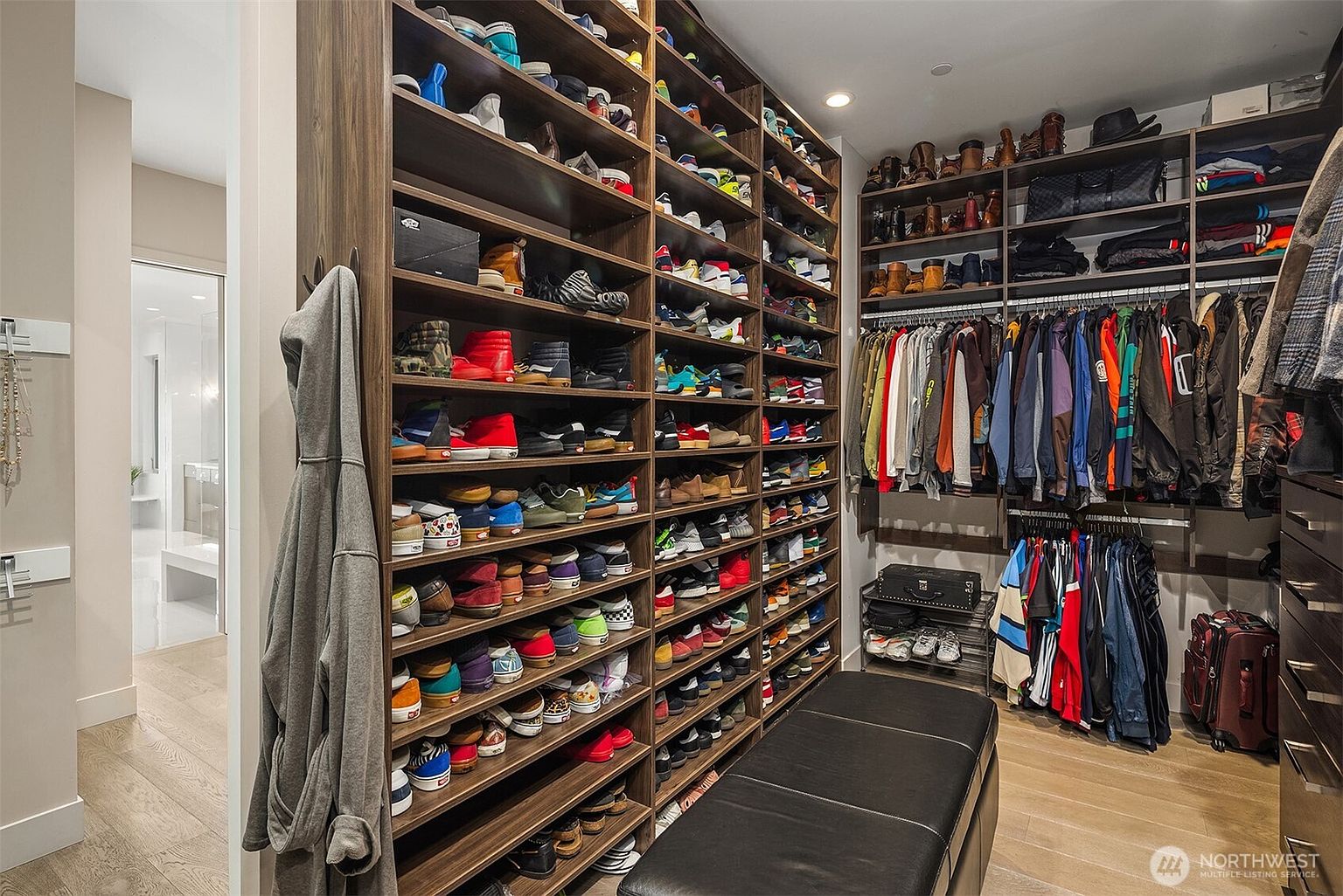 This spacious walk-in closet features extensive custom dark wood shelving units packed with a large collection of footwear and hanging clothing. A long, black leather-upholstered bench sits in the center of the room, providing a functional seating area, while a doorway in the background reveals a glimpse of a bright, modern bathroom. The perspective is from the entrance of the closet, showcasing the organized storage capacity and the transition between the dressing area and the adjacent suite.