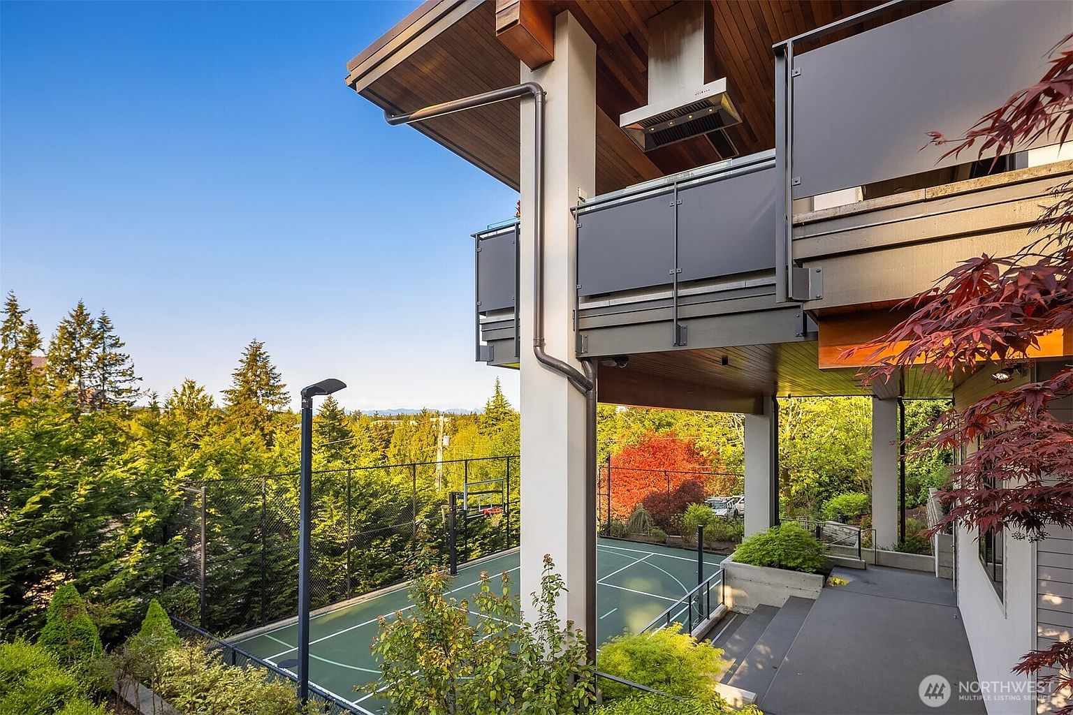 This perspective captures a modern multi-level home featuring a covered outdoor living space with a sleek metal railing and a stainless steel range hood. Below the deck, a private sports court is enclosed by high fencing, surrounded by lush green trees and a vibrant red-leafed tree. The architectural design emphasizes clean lines and a seamless transition between the elevated deck and the recreational area below.