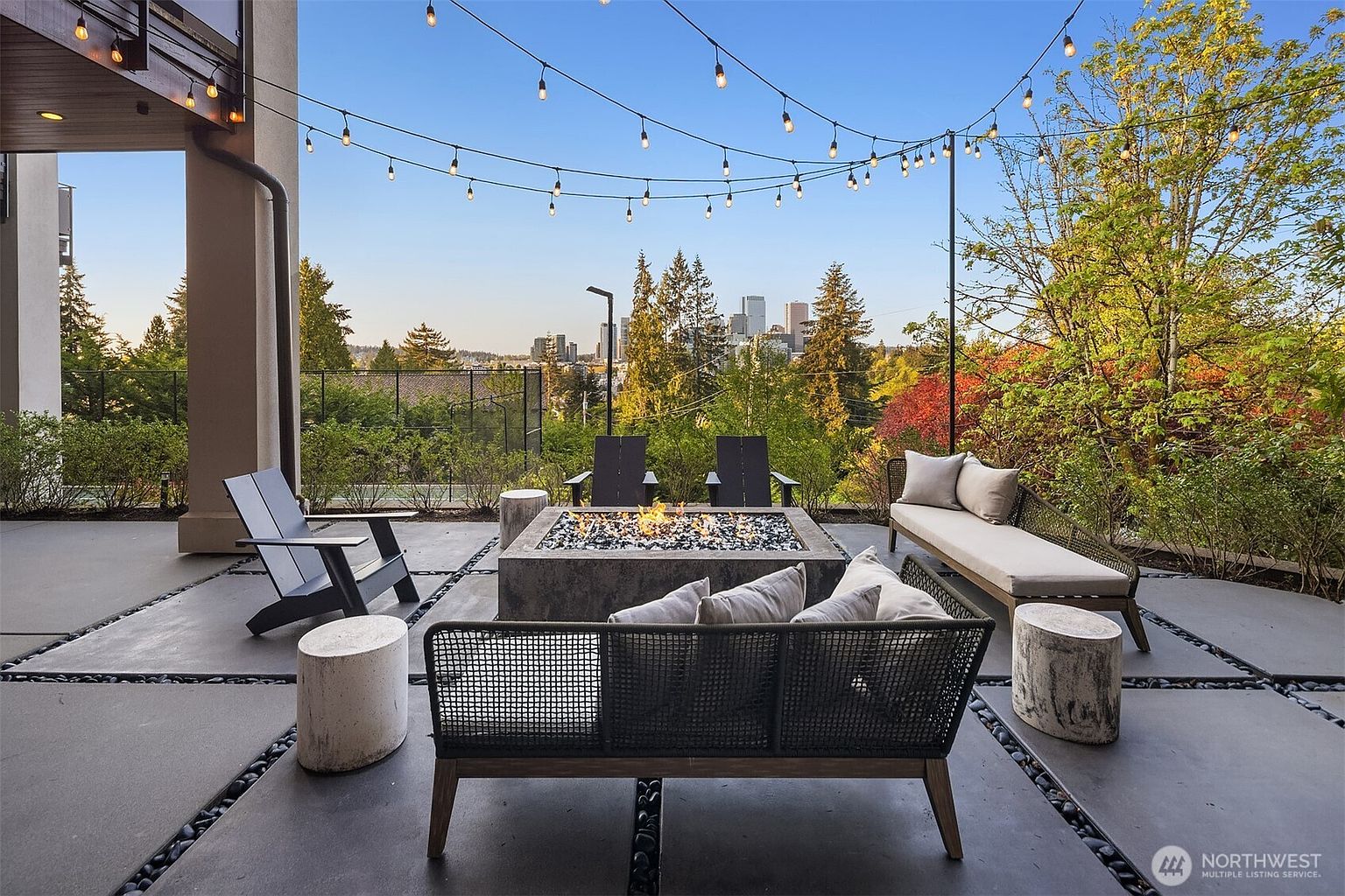 This inviting outdoor patio features a modern concrete fire pit as its centerpiece, surrounded by contemporary lounge seating including Adirondack chairs and a sleek sofa. The space is framed by string lights overhead and offers a stunning, elevated view of the city skyline in the distance. The patio floor is composed of large concrete pavers separated by dark stone accents, creating a clean, structured aesthetic perfect for entertaining.