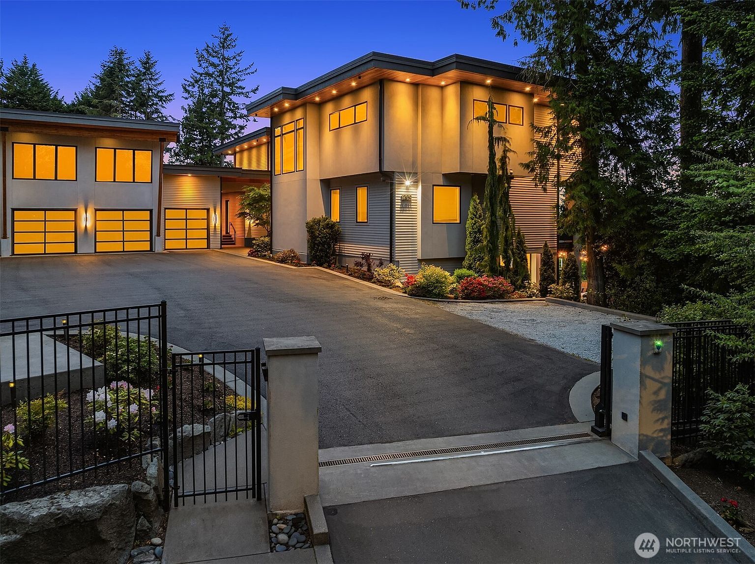 This contemporary multi-level home is captured at dusk, showcasing a warm, inviting glow from its numerous windows and exterior recessed lighting. The property features a spacious paved driveway leading to a three-car garage, with a gated entrance in the foreground and lush, manicured landscaping surrounding the structure. The architectural design emphasizes clean lines and a modern aesthetic, creating a sophisticated and welcoming curb appeal.