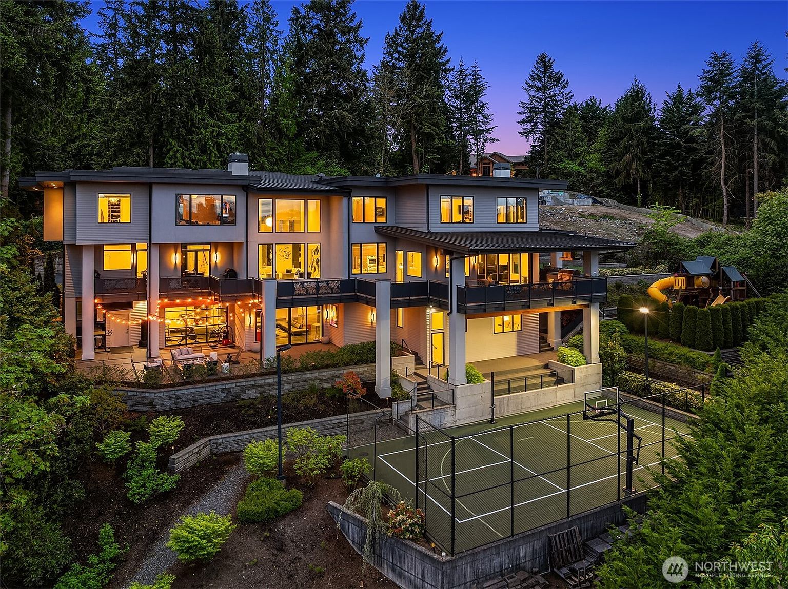 This expansive, multi-level modern residence is captured from an elevated rear perspective, showcasing a sophisticated architectural design with clean lines and large windows. The home features multiple balconies, a covered patio area with string lighting, and a private sports court situated on the lower level. Surrounded by lush, mature trees, the property offers a serene and luxurious atmosphere, perfectly blending indoor-outdoor living spaces.