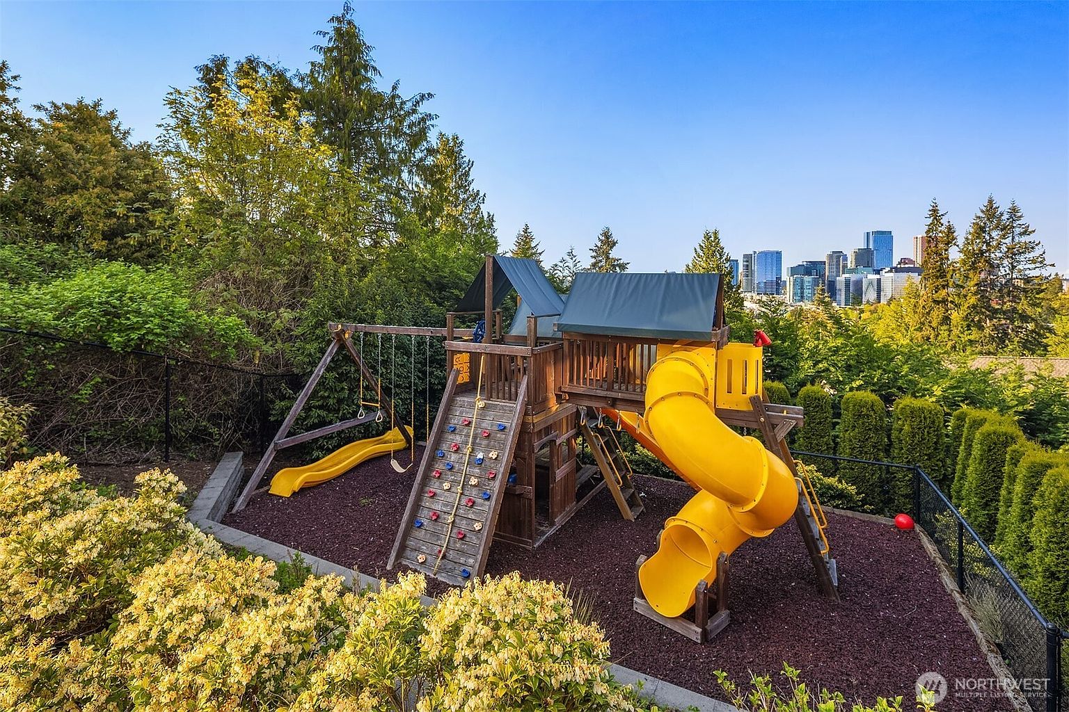 This backyard features a large, multi-level wooden playset equipped with a climbing wall, swings, and a yellow spiral slide, set upon a bed of dark wood mulch. The area is surrounded by lush green trees and manicured hedges, with a clear view of a distant city skyline in the background. The perspective is slightly elevated, capturing the play area as a focal point within a serene, private outdoor setting.