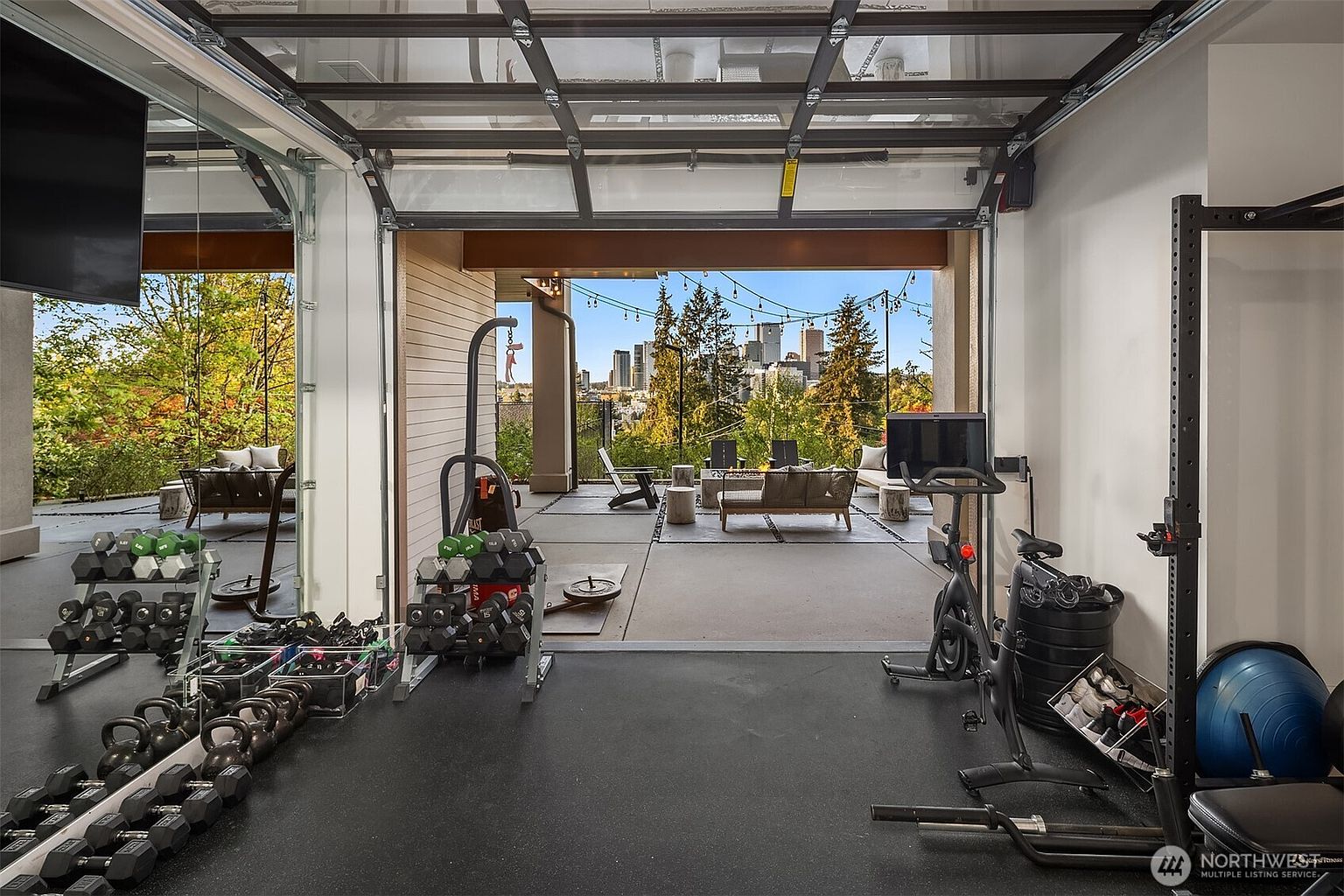 This home gym features a modern, open-concept design with a large, glass-paneled garage door that retracts to seamlessly connect the workout space to an outdoor patio. The gym is equipped with a variety of fitness gear, including a stationary bike, a squat rack, and an extensive collection of dumbbells and kettlebells arranged on racks. Beyond the threshold, the patio offers a lounge area with a fire pit and a stunning, unobstructed view of the city skyline.