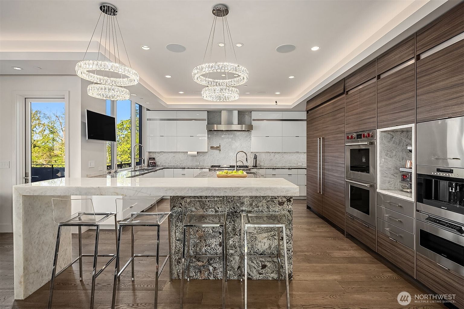 This modern, high-end kitchen features a large central island with a stone waterfall edge and four transparent bar stools. The space is defined by sleek, dark wood-toned cabinetry, white upper cabinets, and integrated stainless steel appliances, including a built-in coffee system. Two ornate, circular crystal chandeliers hang above the island, while large windows to the left provide natural light and a view of the outdoors.