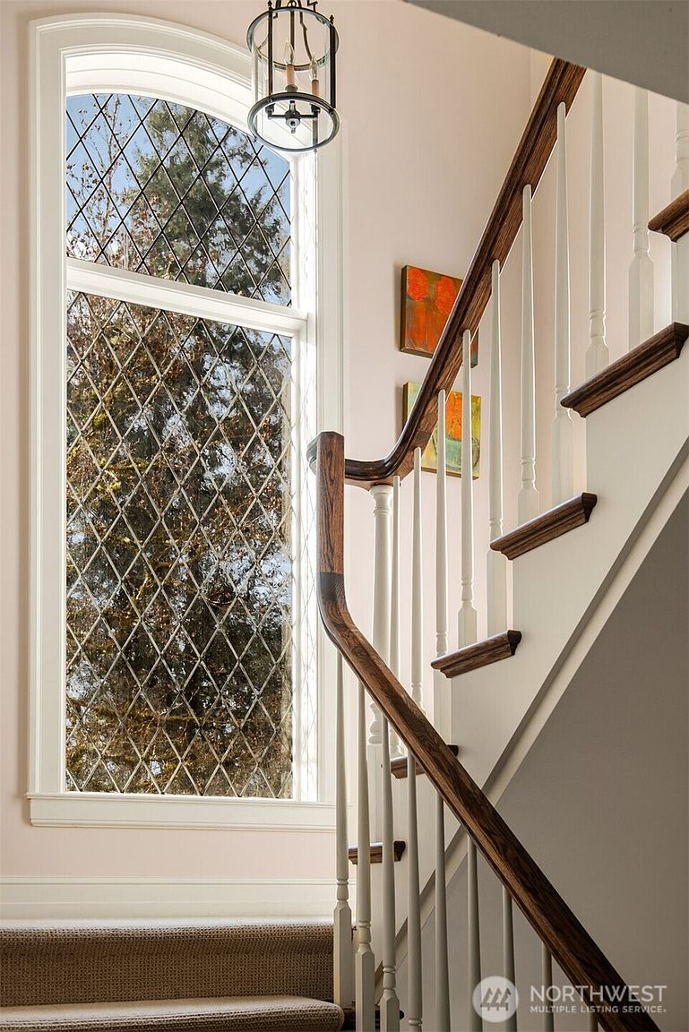 This elegant staircase features a rich, dark wood handrail that contrasts beautifully with the crisp white balusters and neutral-toned carpeted steps. A tall, arched window with a classic diamond-patterned leaded glass design serves as a striking architectural focal point, allowing soft natural light to fill the space. The scene is accented by a sophisticated hanging lantern fixture and framed artwork on the wall, creating a warm and inviting transition area within the home.