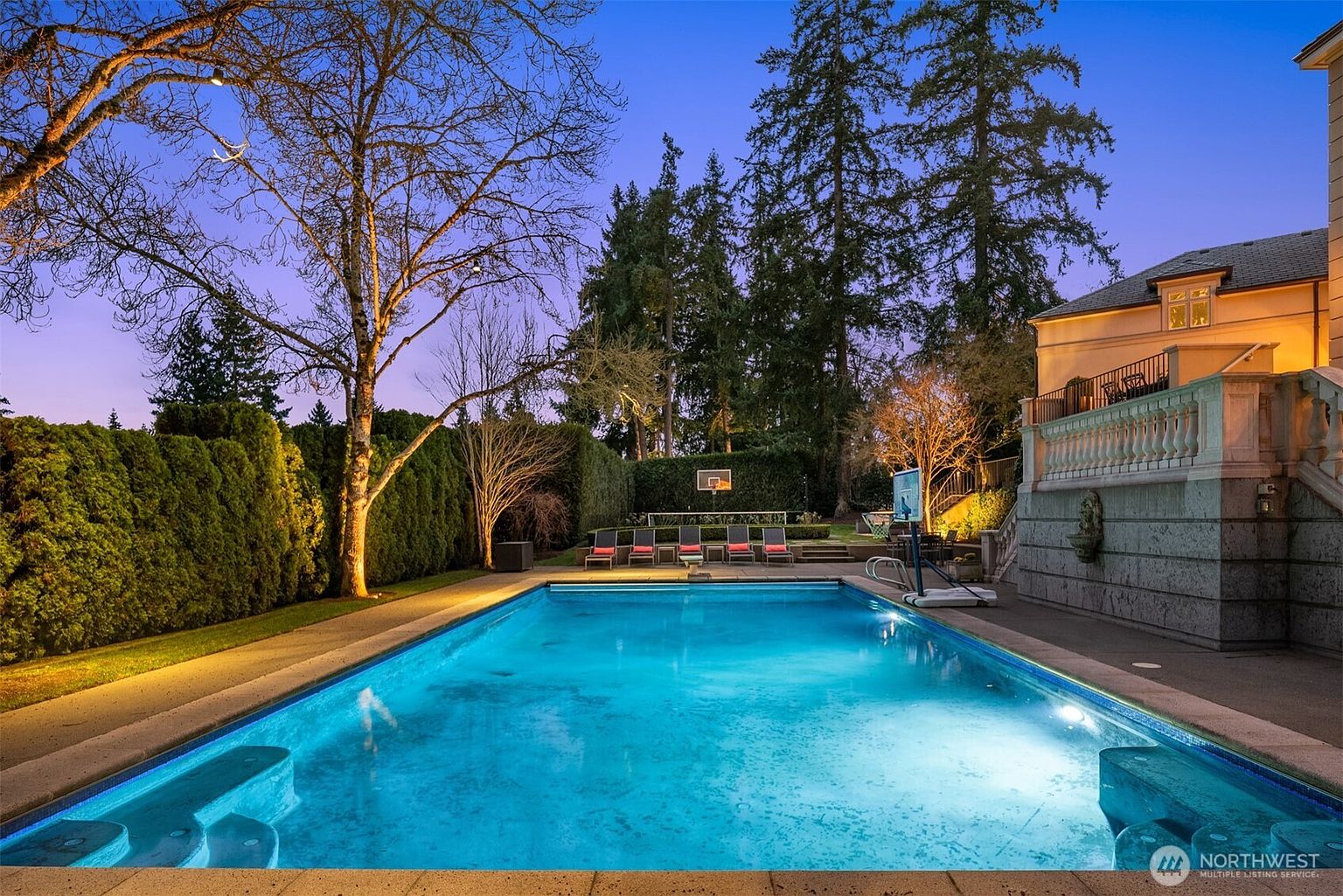 This stunning backyard features a large, illuminated rectangular swimming pool as the centerpiece, set against a backdrop of mature trees and manicured hedges. A stone patio area with lounge chairs and a basketball hoop provides an inviting space for recreation and relaxation, while the adjacent house structure showcases elegant stone masonry and a classic balustrade. The twilight lighting creates a serene and luxurious atmosphere, perfect for evening entertaining.