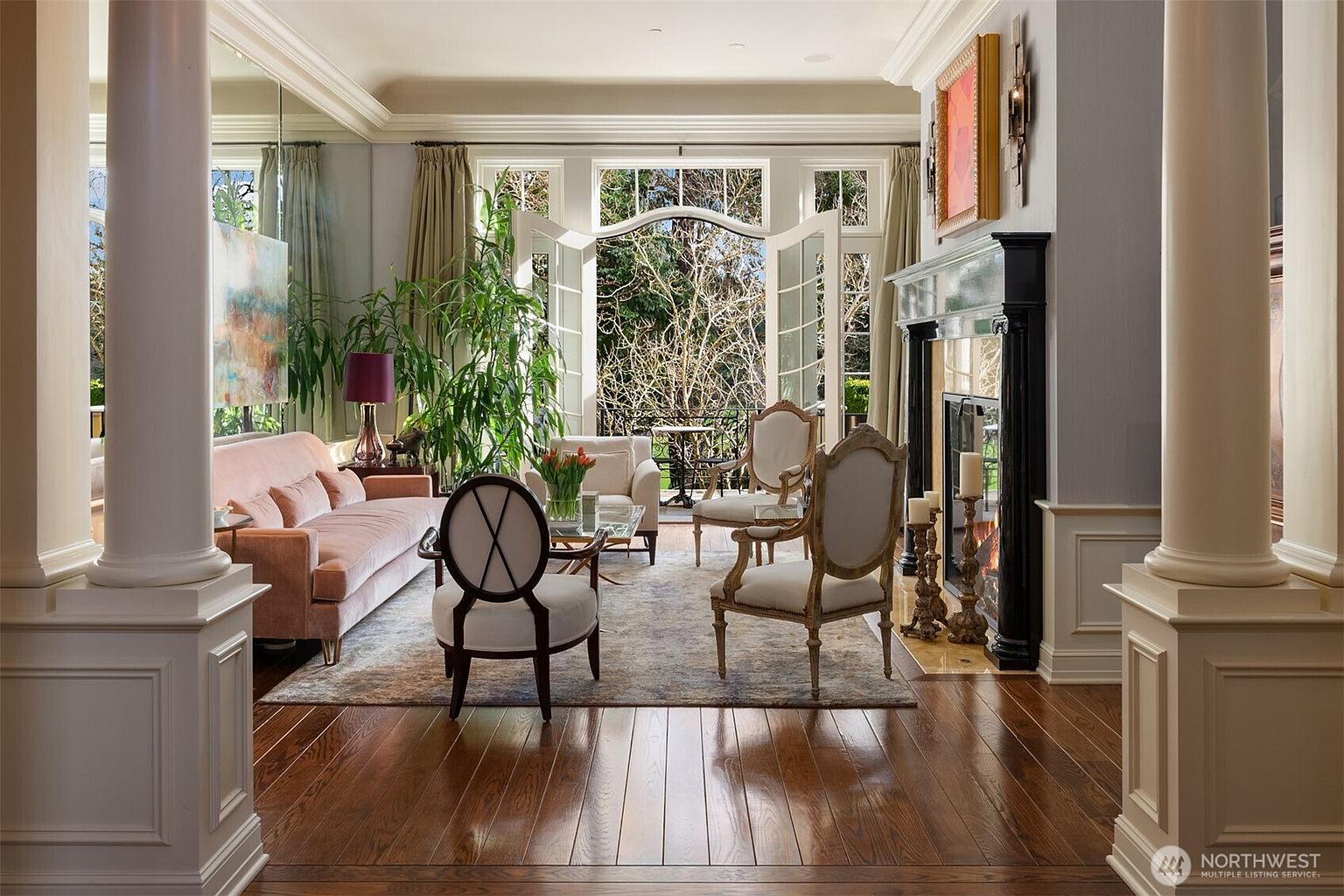 This elegant living room features a sophisticated blend of classic and contemporary design, anchored by a striking black fireplace and rich hardwood flooring. The space is framed by grand white columns and a large mirror, creating an open, airy atmosphere that flows seamlessly through French doors to a private balcony. A plush pink sofa and refined accent chairs are arranged on a textured area rug, offering a luxurious and inviting setting for relaxation.