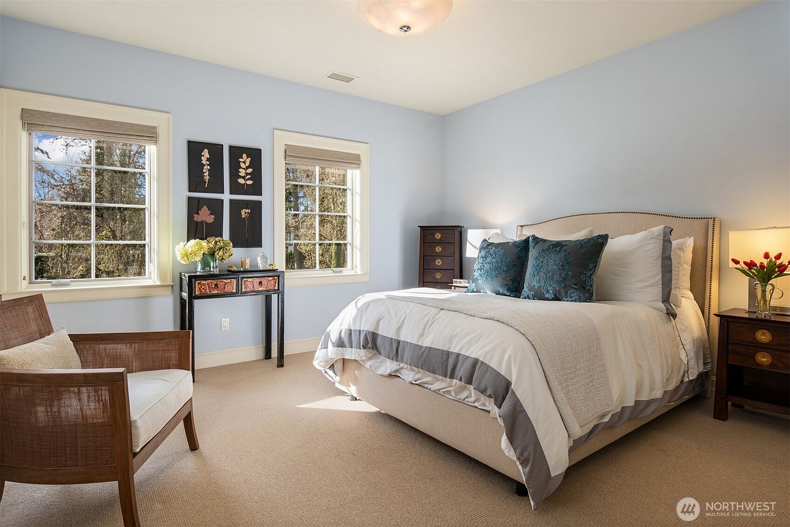 This bright and inviting bedroom features a neutral color palette with soft blue walls and plush beige carpeting. The room is anchored by a queen-sized bed with a tufted headboard, complemented by a wooden nightstand, a matching dresser, and a stylish accent chair. Two large windows allow for ample natural light, while a console table adorned with framed botanical art adds a touch of elegance to the space.