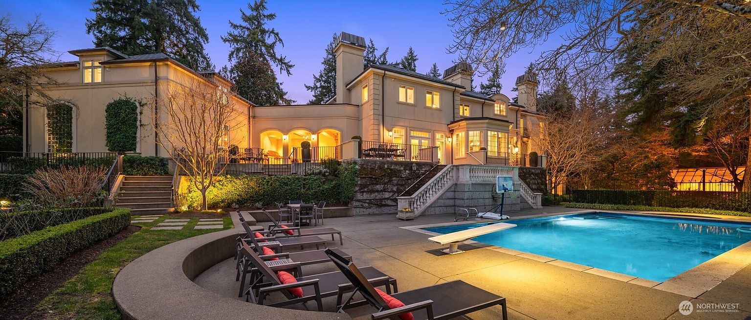 This stunning rear exterior view of a grand, multi-story estate showcases a luxurious swimming pool and expansive stone patio area at twilight. The home features elegant architectural details, including arched loggias, stone staircases, and warm, inviting lighting that highlights the manicured landscaping. The scene captures a sophisticated and serene atmosphere, perfect for high-end outdoor entertaining.