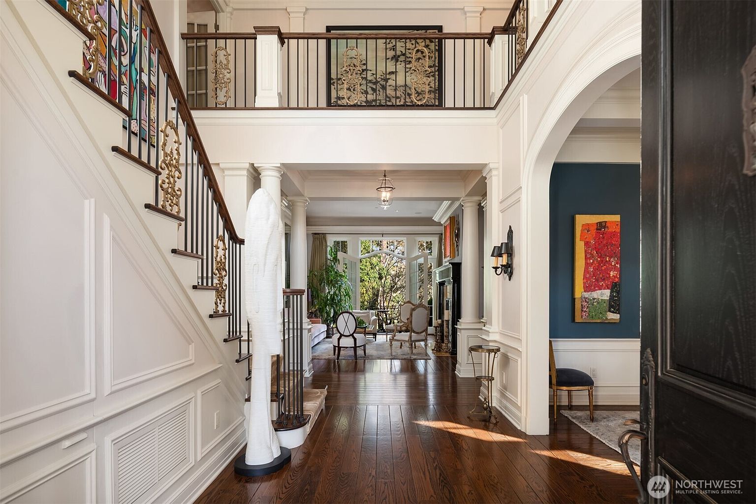 This grand entryway features a dramatic staircase with ornate metal railings and a striking white sculptural piece, leading into a bright, open-concept living space. The foyer boasts rich hardwood floors, elegant wainscoting, and a high-ceilinged landing with decorative ironwork, creating a sophisticated and welcoming atmosphere. The perspective looks through the foyer toward a sunlit living area, highlighting the home's spacious and luxurious architectural design.