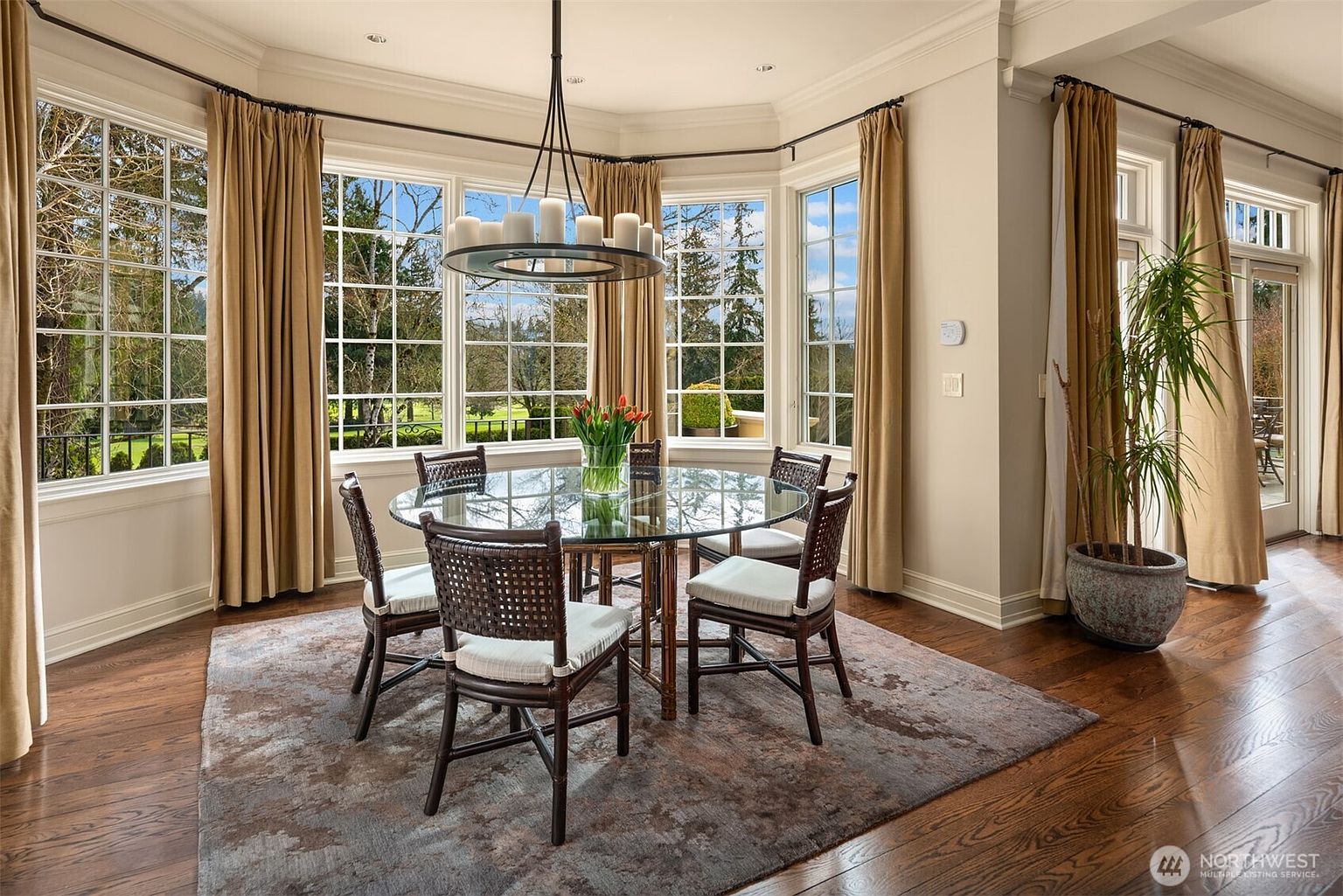 This elegant dining room features a circular glass-top table surrounded by four dark wicker chairs, set atop a textured area rug on rich hardwood floors. A large, multi-light chandelier hangs above the table, while expansive bay windows draped in floor-to-ceiling curtains provide abundant natural light and views of the lush outdoor landscape. The space exudes a sophisticated, airy atmosphere, perfect for formal or casual dining.
