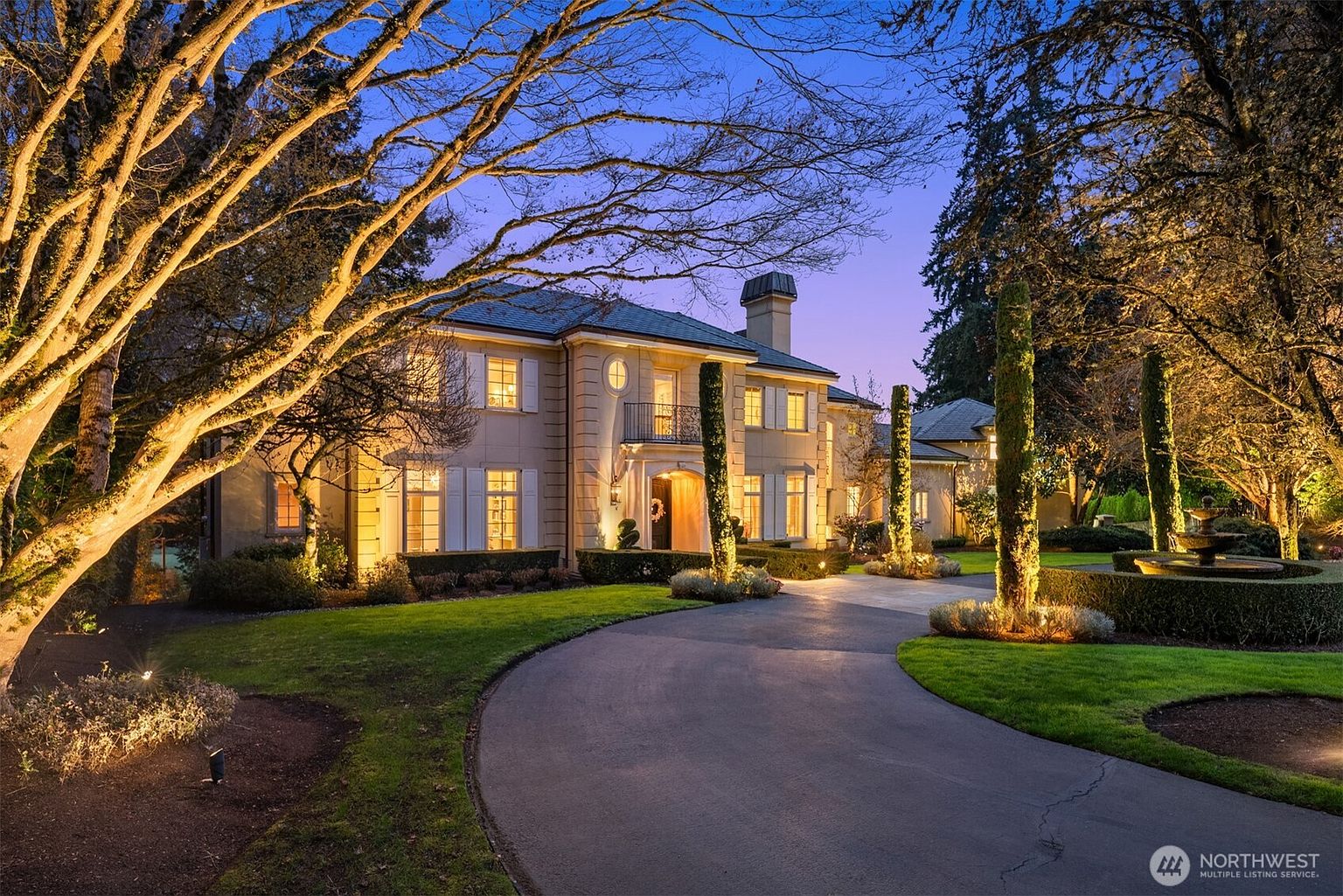 This elegant, two-story estate is captured at twilight, showcasing a grand facade with symmetrical architecture, white shutters, and a prominent arched entryway. A sweeping paved driveway leads to the home, framed by manicured lawns, mature trees, and decorative landscaping illuminated by warm, inviting exterior lighting. The overall impression is one of luxury, sophistication, and curb appeal, perfect for a high-end real estate presentation.