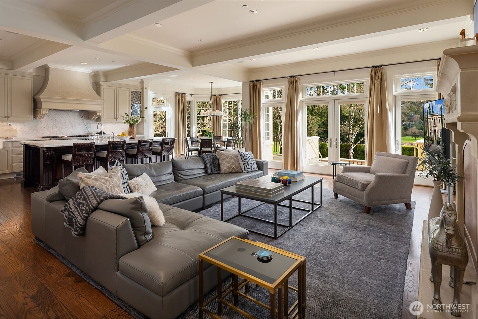 This expansive, open-concept living room features a large, plush grey sectional sofa and a matching armchair, centered around a modern black-framed coffee table on a textured area rug. The space flows seamlessly into a gourmet kitchen with a dark wood island and marble countertops, while floor-to-ceiling windows and French doors flood the room with natural light and offer views of the lush backyard. The high ceilings and neutral color palette create an elegant, airy, and sophisticated atmosphere perfect for entertaining.