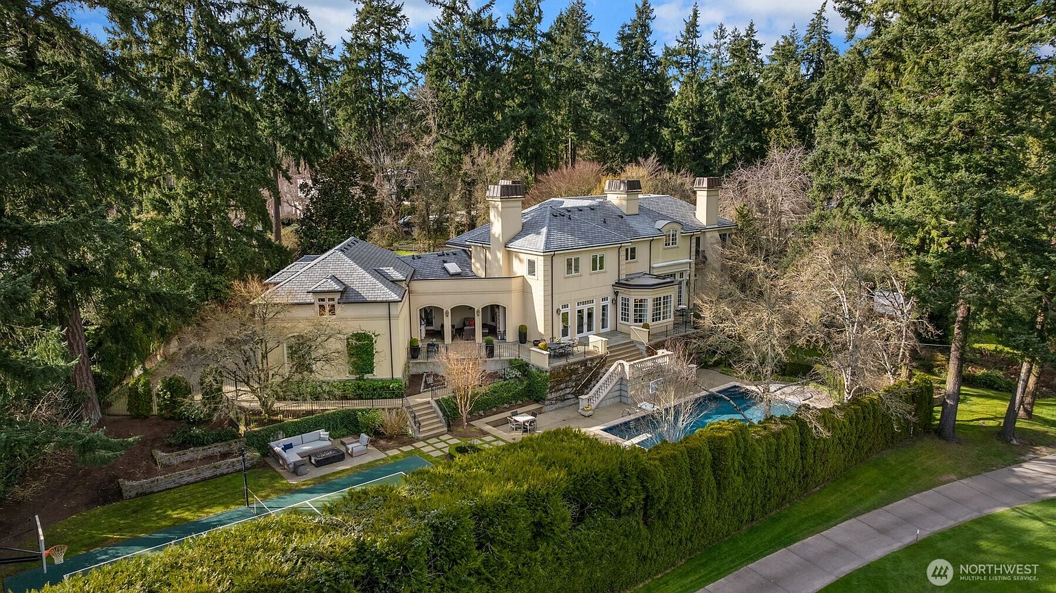 This stunning aerial view captures a grand, multi-story estate nestled within a lush, mature forest. The property features a sophisticated architectural design with a slate roof, a sprawling rear terrace, and a private swimming pool set against a backdrop of towering evergreen trees. The well-manicured grounds include a tennis court and elegant landscaping, creating a serene and luxurious atmosphere.
