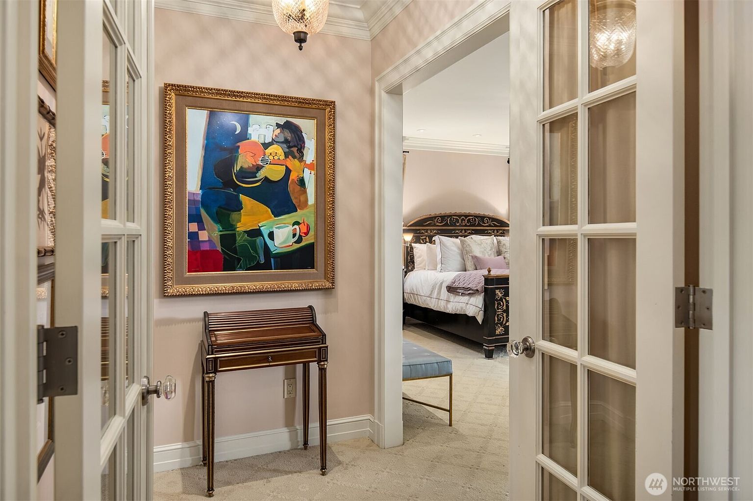 This image captures a view through a set of glass-paned French doors into a sophisticated bedroom suite. In the foreground, a small wooden writing desk sits against a wall adorned with a vibrant, abstract painting, creating a refined transitional space. The perspective draws the eye from the neutral-toned hallway directly into the primary bedroom, highlighting the elegant decor and seamless flow between rooms.
