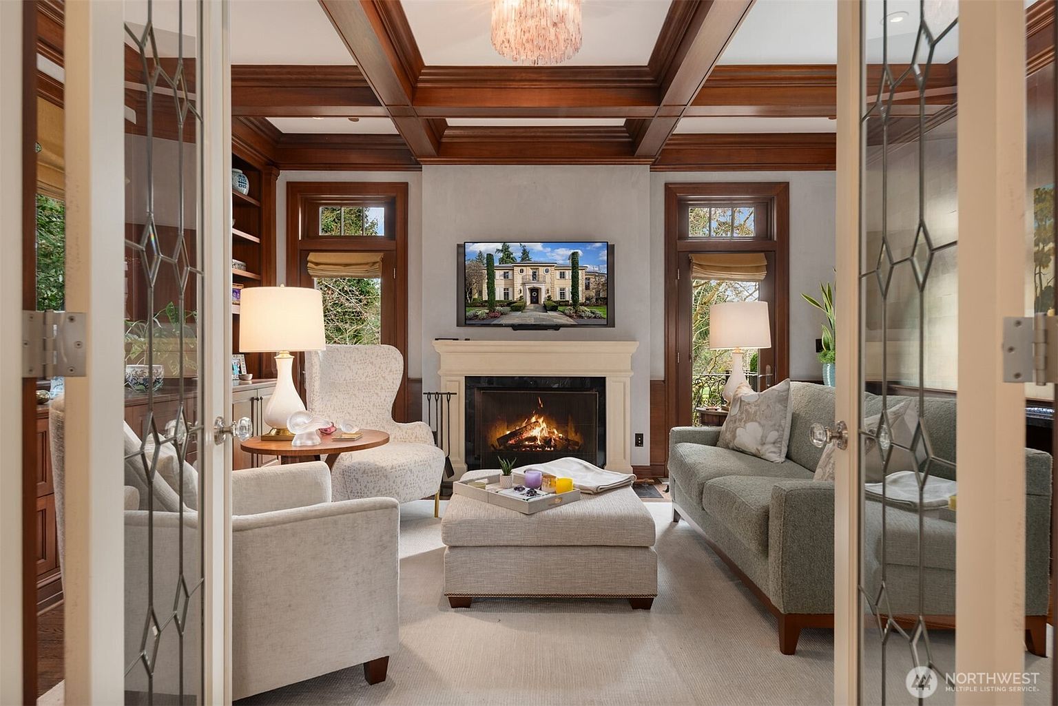 This elegant living room is captured from the perspective of looking through glass-paned French doors, creating a welcoming and sophisticated atmosphere. The space features a coffered wood ceiling, a cozy fireplace with a mounted television, and plush, neutral-toned seating arranged around a central ottoman. Rich wood trim and built-in shelving add a sense of warmth and luxury to this well-appointed sitting area.