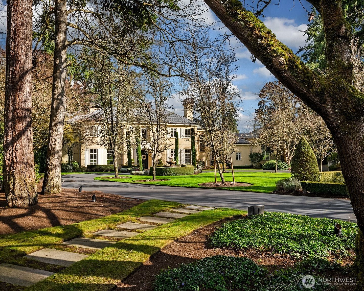 This grand, two-story estate features a classic architectural style with a symmetrical facade, light-colored stucco, and dark-framed windows. The property is approached by a sweeping paved driveway that leads to a circular fountain centerpiece, all set within a lush, manicured lawn surrounded by mature trees. The perspective is captured from a low angle, emphasizing the stately presence of the home and the expansive, park-like grounds.