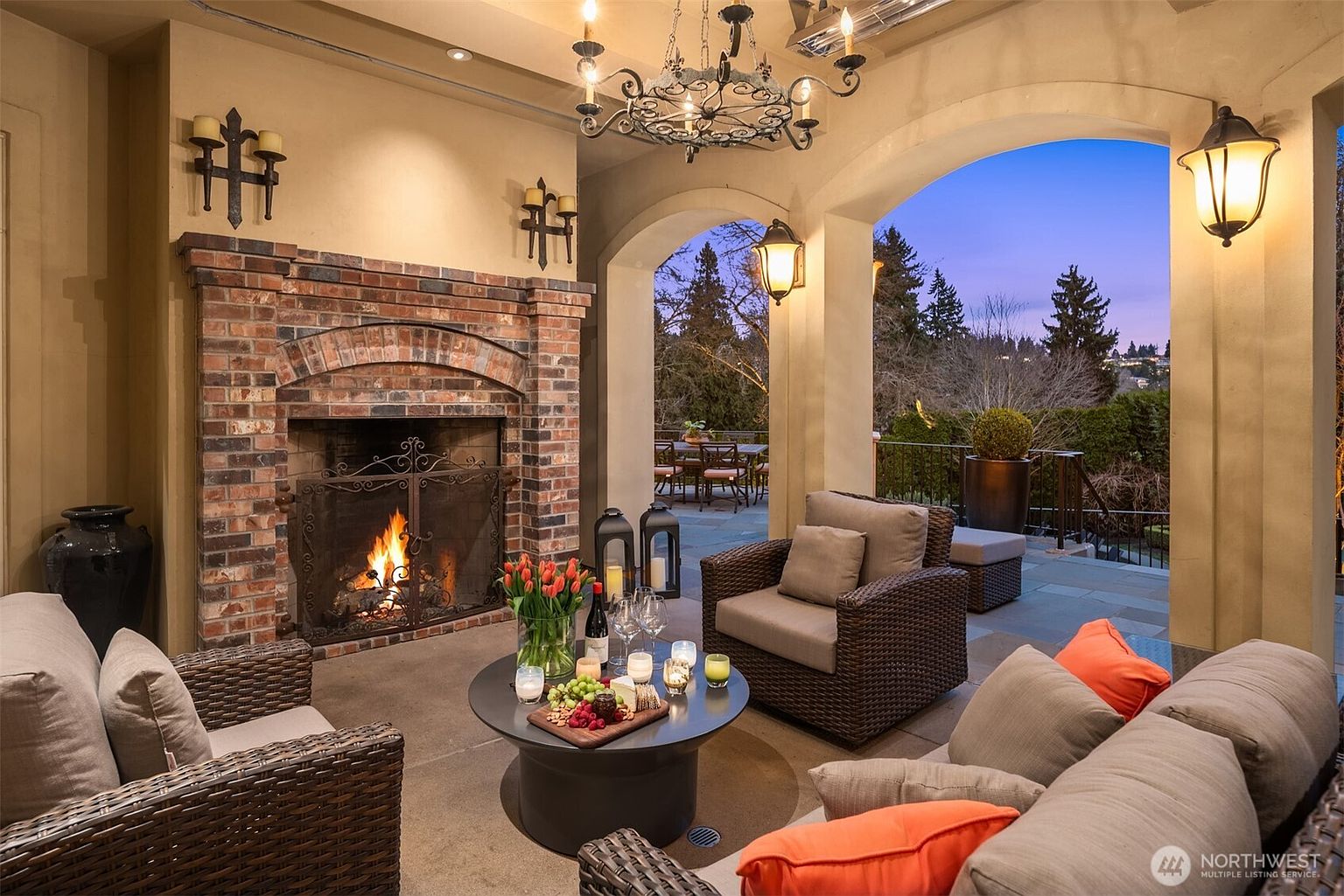 This luxurious covered patio features a grand brick fireplace as its centerpiece, creating a warm and inviting atmosphere for outdoor entertaining. The space is furnished with comfortable wicker seating, a central coffee table with refreshments, and elegant lighting, all overlooking a scenic, tree-lined landscape at dusk. The architectural design blends classic masonry with an open-air layout, offering a sophisticated and tranquil retreat.