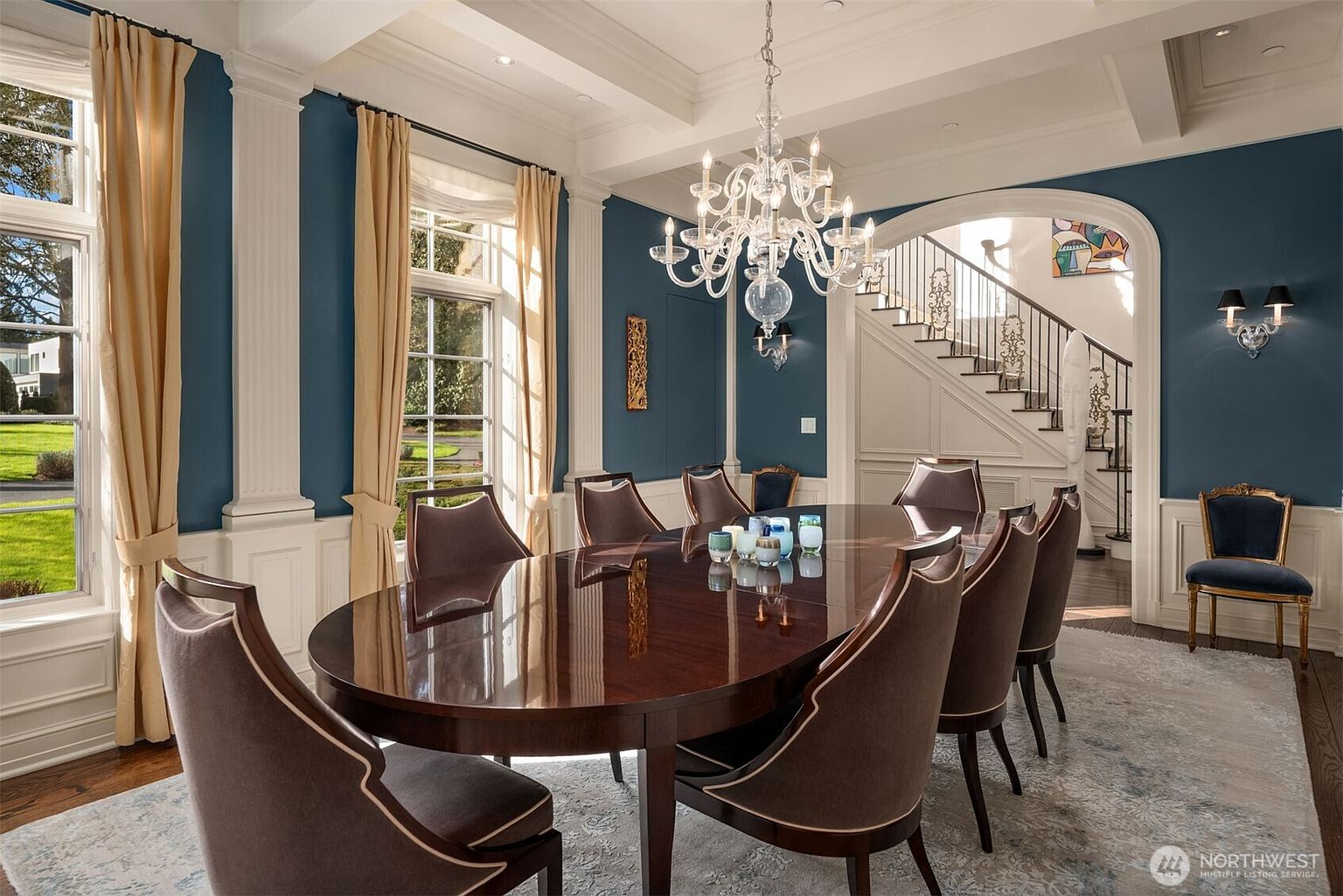 This elegant formal dining room features a large, polished mahogany table surrounded by upholstered chairs, set beneath a sophisticated crystal chandelier. The room is defined by deep blue walls, crisp white wainscoting, and tall windows draped in soft gold curtains, creating a refined and luxurious atmosphere. An arched entryway leads to a staircase with ornate iron railings, adding a sense of architectural grandeur to the space.