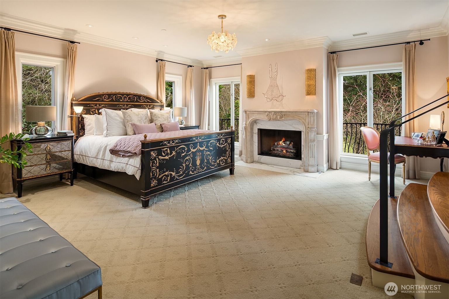 This elegant primary bedroom features a sophisticated, ornate black and gold bed frame as the centerpiece, complemented by plush, neutral carpeting and soft pink walls. The room is bathed in natural light from multiple large windows and includes a cozy fireplace with a marble mantel, a dedicated vanity area, and a crystal chandelier that adds a touch of luxury. The overall aesthetic is refined and opulent, offering a serene and high-end retreat.