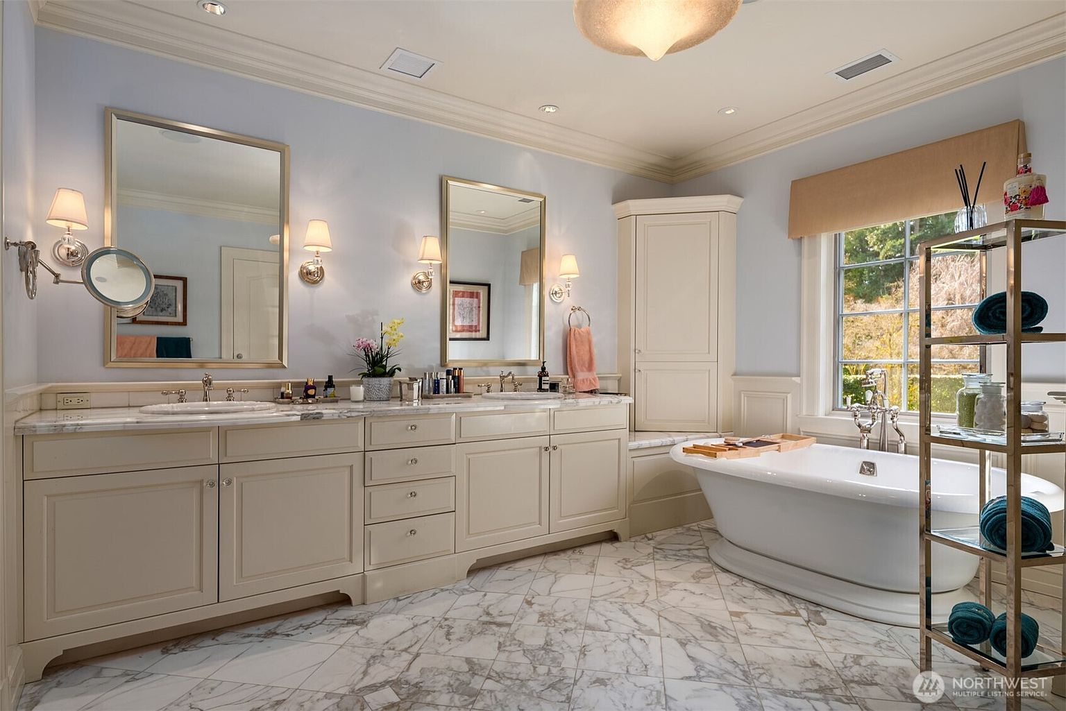 This elegant primary bathroom features a double vanity with marble countertops, dual mirrors, and classic cream-colored cabinetry. A freestanding soaking tub sits beneath a large window, complemented by a corner storage unit and a tall, open-shelving metal rack. The room is finished with luxurious marble flooring and soft blue walls, creating a serene and sophisticated spa-like atmosphere.