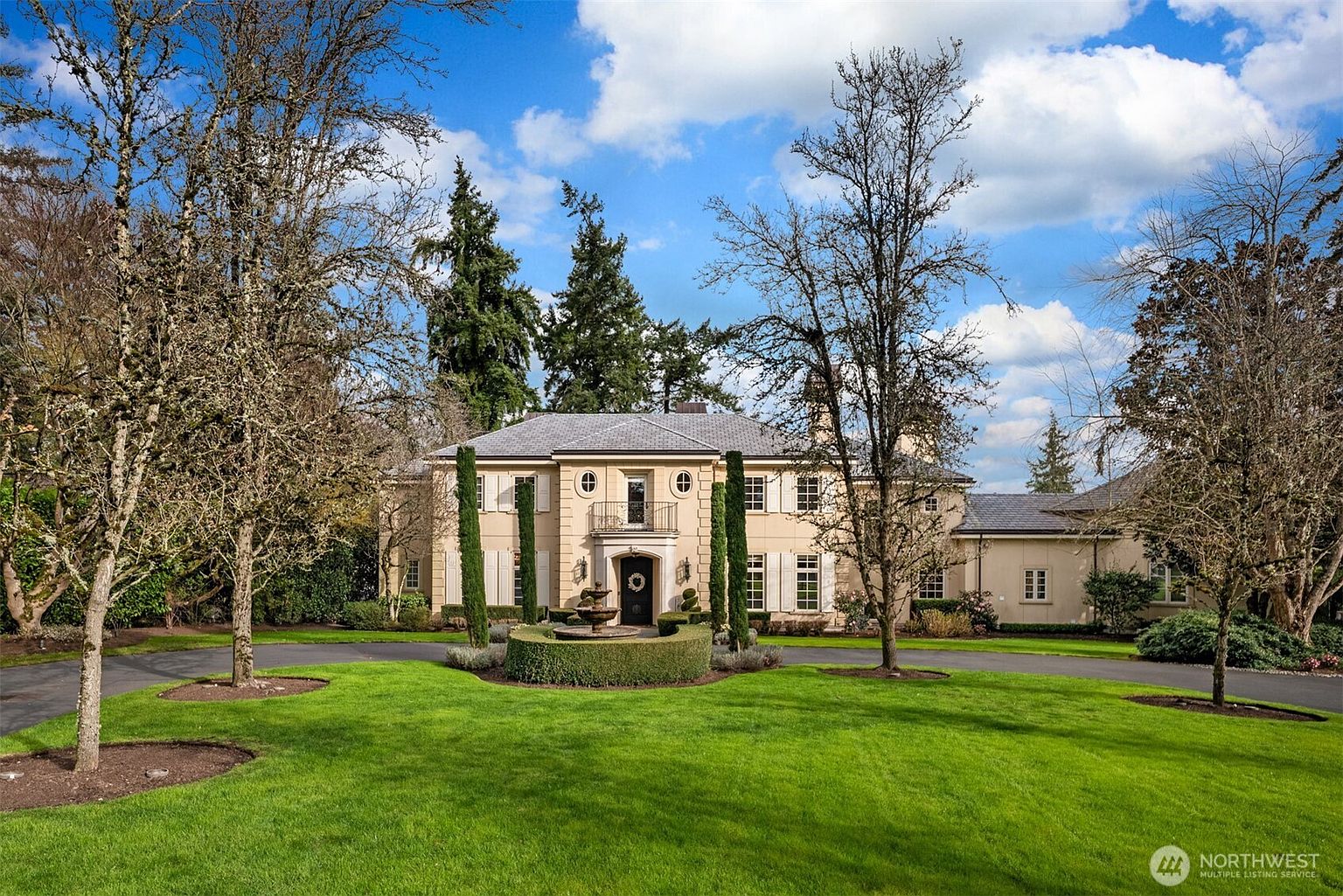This grand, two-story estate features a symmetrical, classic architectural design with a light-colored stucco facade and a dark shingled roof. A circular driveway surrounds a central fountain and manicured landscaping, creating an elegant and welcoming approach to the main entrance. The property is framed by mature trees and lush green lawns, conveying a sense of luxury, privacy, and timeless sophistication.