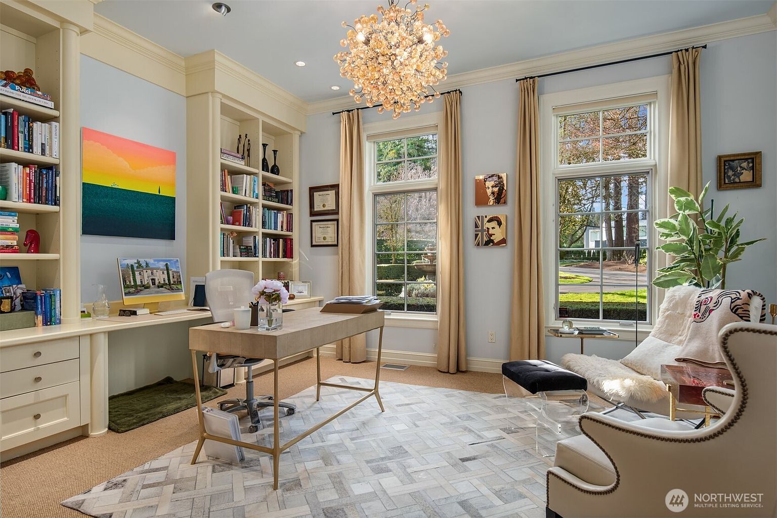 This elegant home office features built-in floor-to-ceiling bookshelves, a sleek desk, and a comfortable reading nook with a plush armchair. Large windows with floor-length curtains flood the room with natural light, highlighting the sophisticated decor and the intricate geometric area rug. The space is anchored by a stunning, multi-bulb chandelier, creating a refined and productive atmosphere.