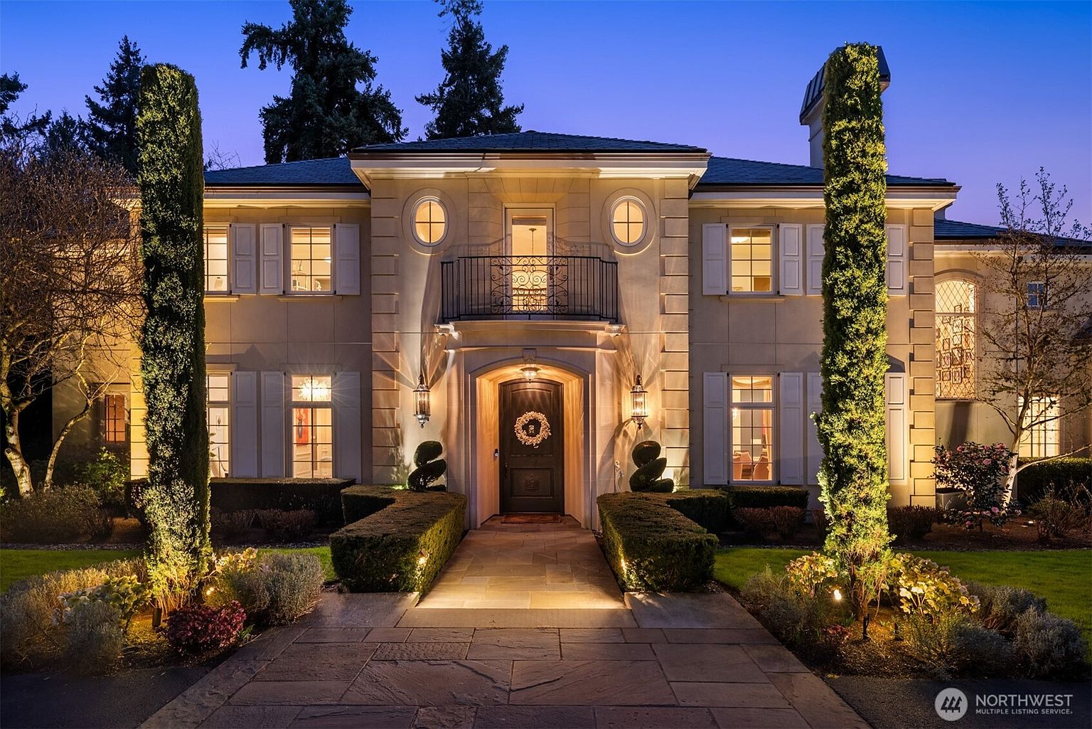 This elegant, symmetrical two-story estate features a classic Mediterranean-inspired facade with a stone exterior, arched entryway, and a decorative wrought-iron balcony. The home is framed by two tall, manicured cypress trees and illuminated by warm, inviting landscape lighting that highlights the architectural details at dusk. A stone pathway leads directly to the grand front door, creating a sophisticated and welcoming curb appeal.