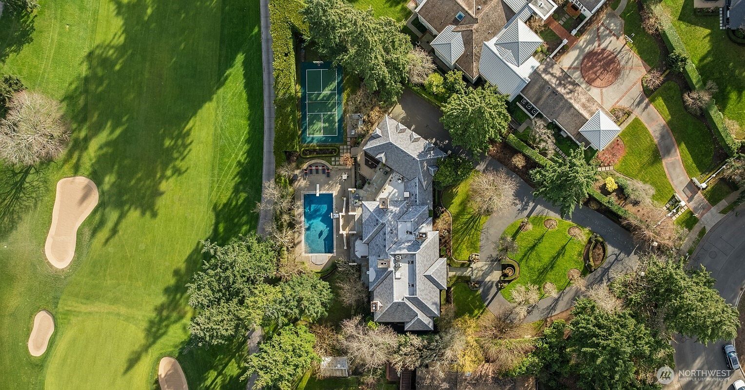 This high-angle aerial view captures a sprawling luxury estate situated directly adjacent to a lush green golf course. The property features a large, multi-gabled residence, a private tennis court, and a rectangular swimming pool surrounded by a stone patio. The grounds are meticulously landscaped with mature trees, expansive lawns, and a circular driveway, creating an impression of exclusivity and refined living.