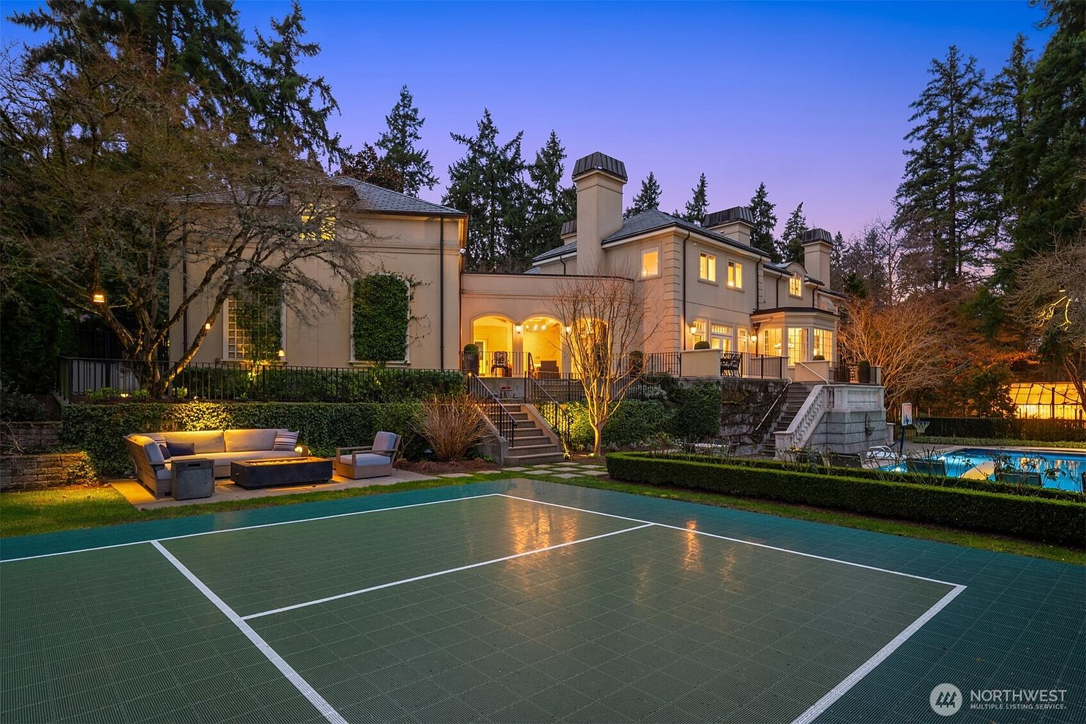This elegant estate features a sprawling rear view at dusk, showcasing a multi-level terrace with a cozy fire pit seating area and a pristine swimming pool. The classic architectural design is complemented by manicured hedges, a private sports court, and warm exterior lighting that highlights the home's sophisticated facade. The perspective captures the seamless transition between the luxurious indoor living spaces and the expansive, well-appointed outdoor recreational grounds.