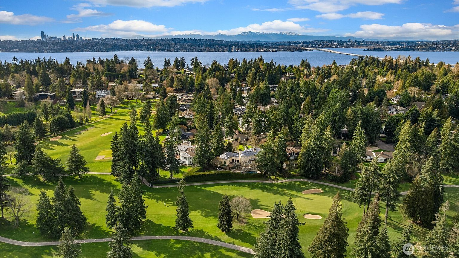 This high-angle aerial view captures a sprawling residential property nestled directly adjacent to a lush, green golf course. The scene features a large, traditional-style home surrounded by dense, mature evergreen trees, with a scenic backdrop of a wide lake, a bridge, and distant snow-capped mountains under a bright, cloudy sky. The perspective emphasizes the expansive, tranquil setting and the prime location of the home within a verdant, park-like community.