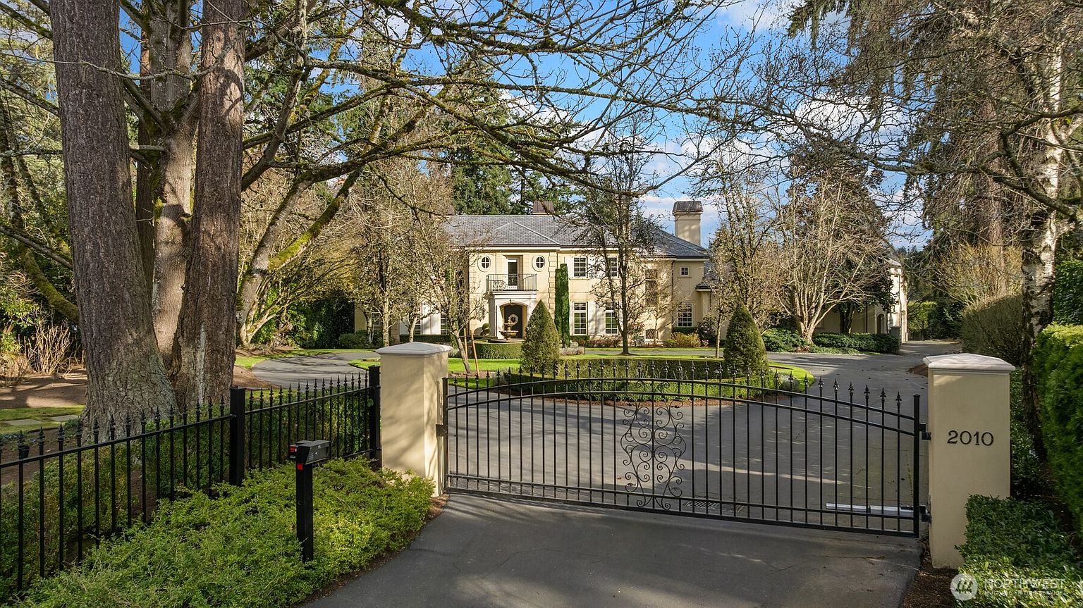 This grand, stately residence is viewed from behind a secure, gated entrance, emphasizing its privacy and exclusivity. The home features classic architectural elements, a circular driveway, and a lush, mature landscape with towering trees that frame the property. The perspective captures the impressive scale and elegant curb appeal of this estate, suggesting a luxurious and secluded lifestyle.