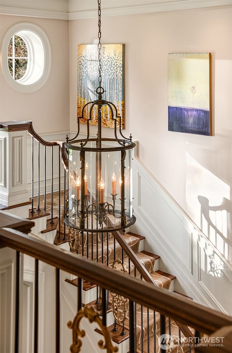 This elegant staircase features a classic design with a rich wooden handrail, dark metal balusters, and a patterned runner carpet. A grand, cylindrical lantern-style chandelier hangs as a focal point, while the walls are adorned with two pieces of contemporary art and a circular window. The perspective is captured from the upper landing, looking down the staircase to emphasize the architectural depth and sophisticated interior aesthetic.