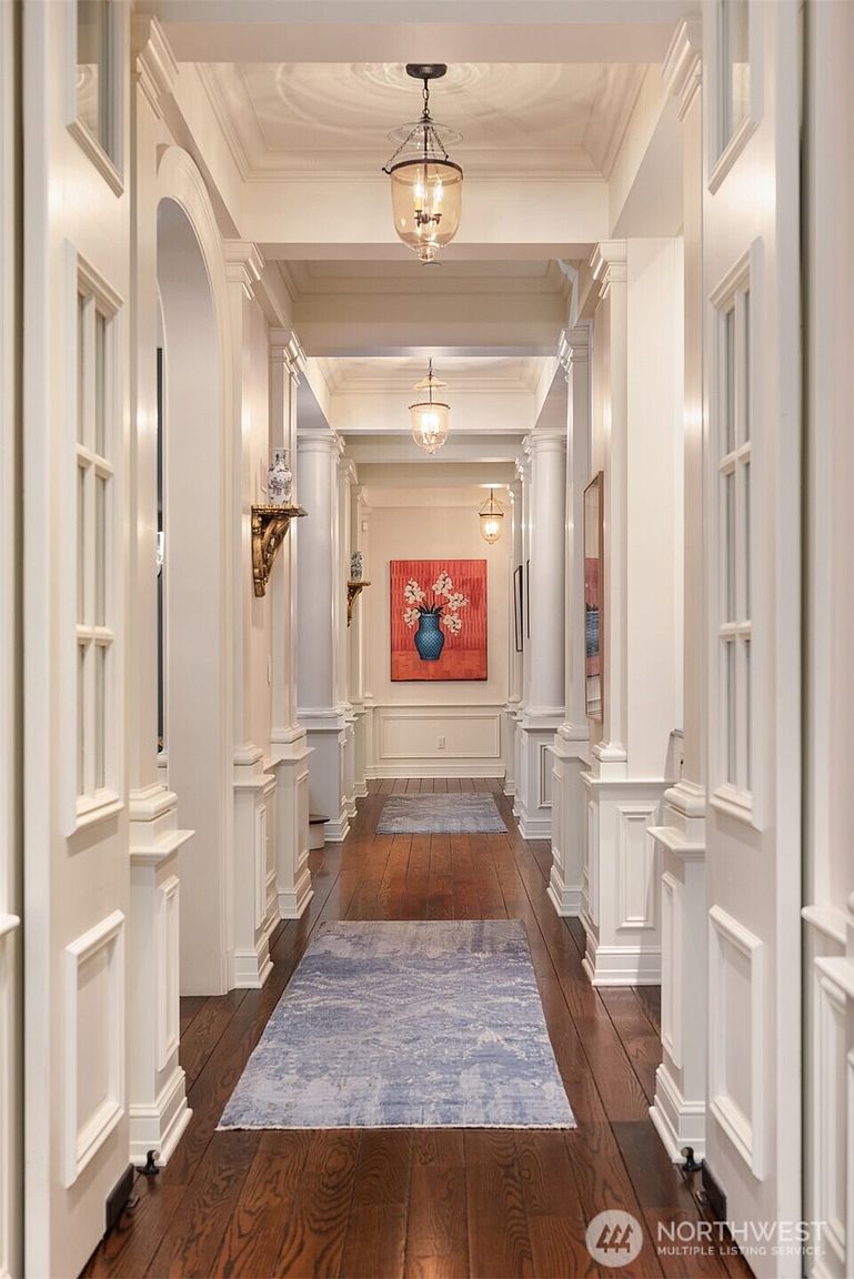 This elegant, long hallway features rich hardwood flooring, classic white wainscoting, and architectural columns that create a sense of depth and grandeur. The space is illuminated by a series of sophisticated glass pendant lights and adorned with a vibrant piece of wall art at the far end. The perspective is a centered, eye-level shot that emphasizes the symmetrical design and refined, traditional aesthetic of the home.
