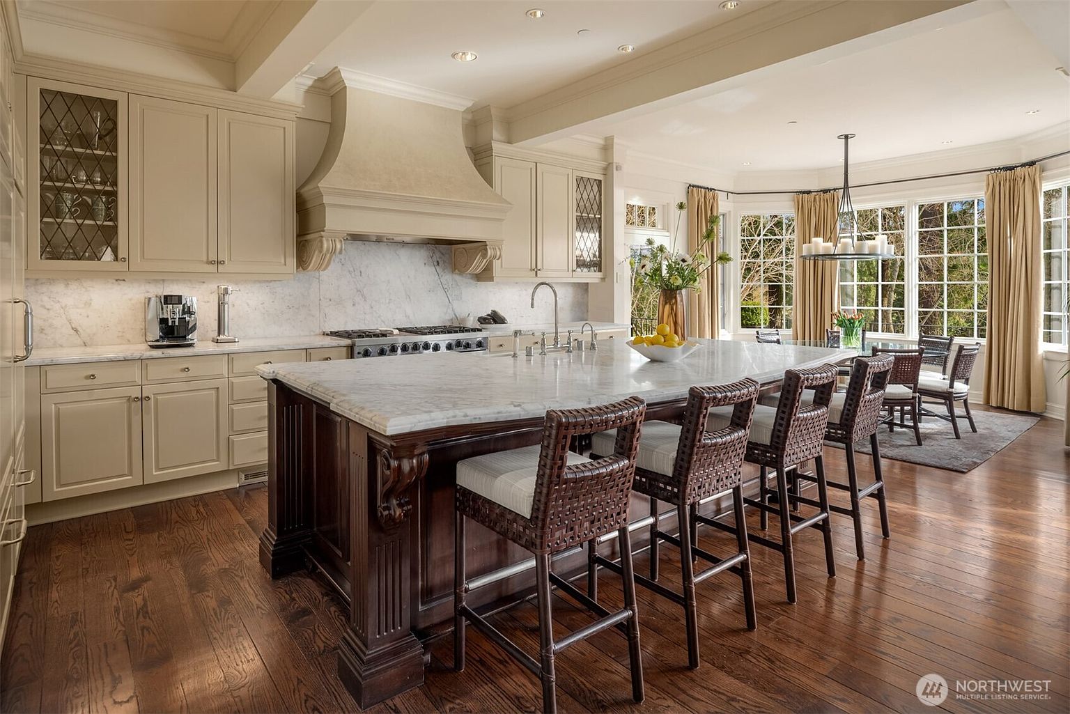 This elegant, traditional-style kitchen features a large, dark wood center island with a marble countertop and four woven bar stools. The space is anchored by light-colored cabinetry, a custom range hood, and a marble backsplash, all illuminated by recessed lighting and a chandelier over the adjacent dining area. Large windows with floor-to-ceiling curtains provide ample natural light and a view of the outdoors, creating a warm and inviting atmosphere.
