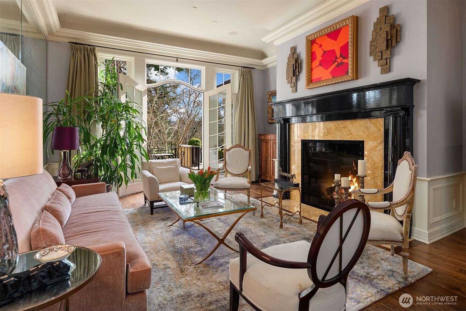 This elegant living room features a sophisticated blend of traditional and contemporary design, anchored by a striking black marble fireplace with a warm, glowing fire. The space is furnished with a plush velvet sofa, classic upholstered armchairs, and a sleek glass-topped coffee table, all arranged on a textured area rug. Large French doors open to a scenic outdoor view, flooding the room with natural light and creating a seamless transition between the refined interior and the lush landscape.