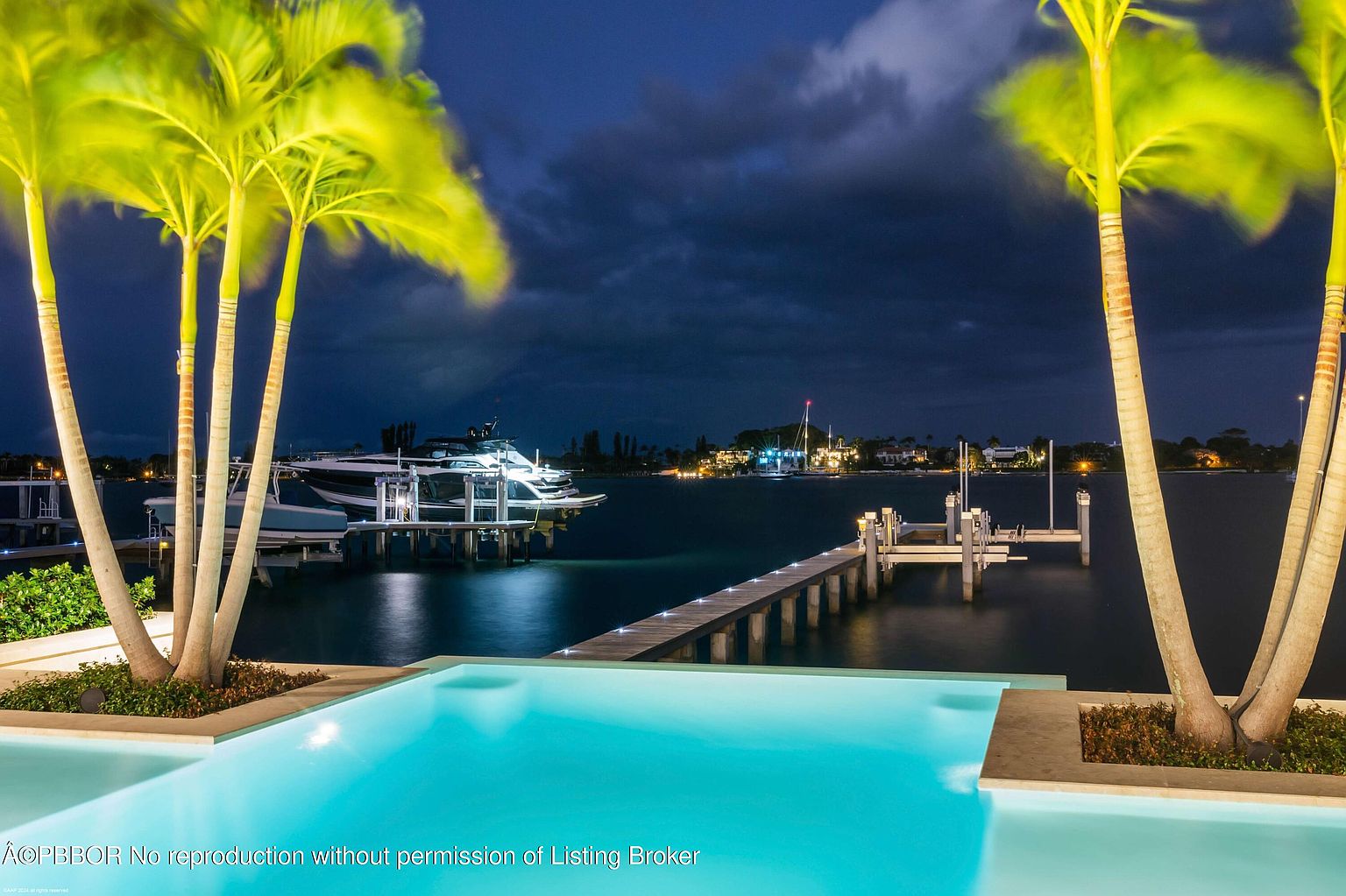 This image showcases a luxurious outdoor pool area at night, framed by palm trees. The pool's turquoise water reflects the ambient light, creating a serene atmosphere. In the background, a waterfront view includes a dock and boats, suggesting a high-end coastal property.