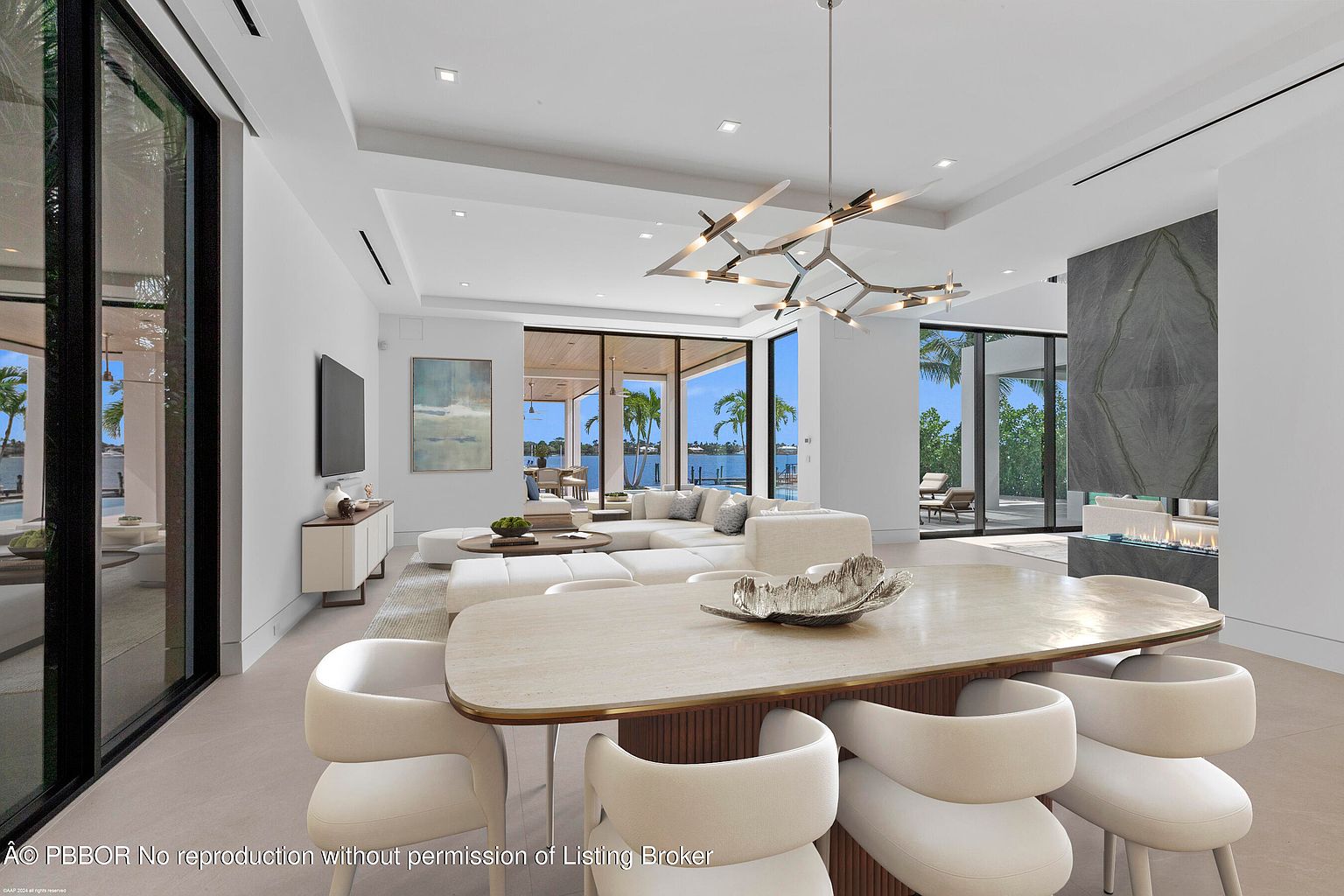 This is a bright and airy living room with a modern design. The room features a large white sectional sofa, a dining table with white chairs, and a striking chandelier. Large windows offer views of the water and palm trees, creating a seamless indoor-outdoor living experience. A fireplace with a stone surround adds a touch of elegance.